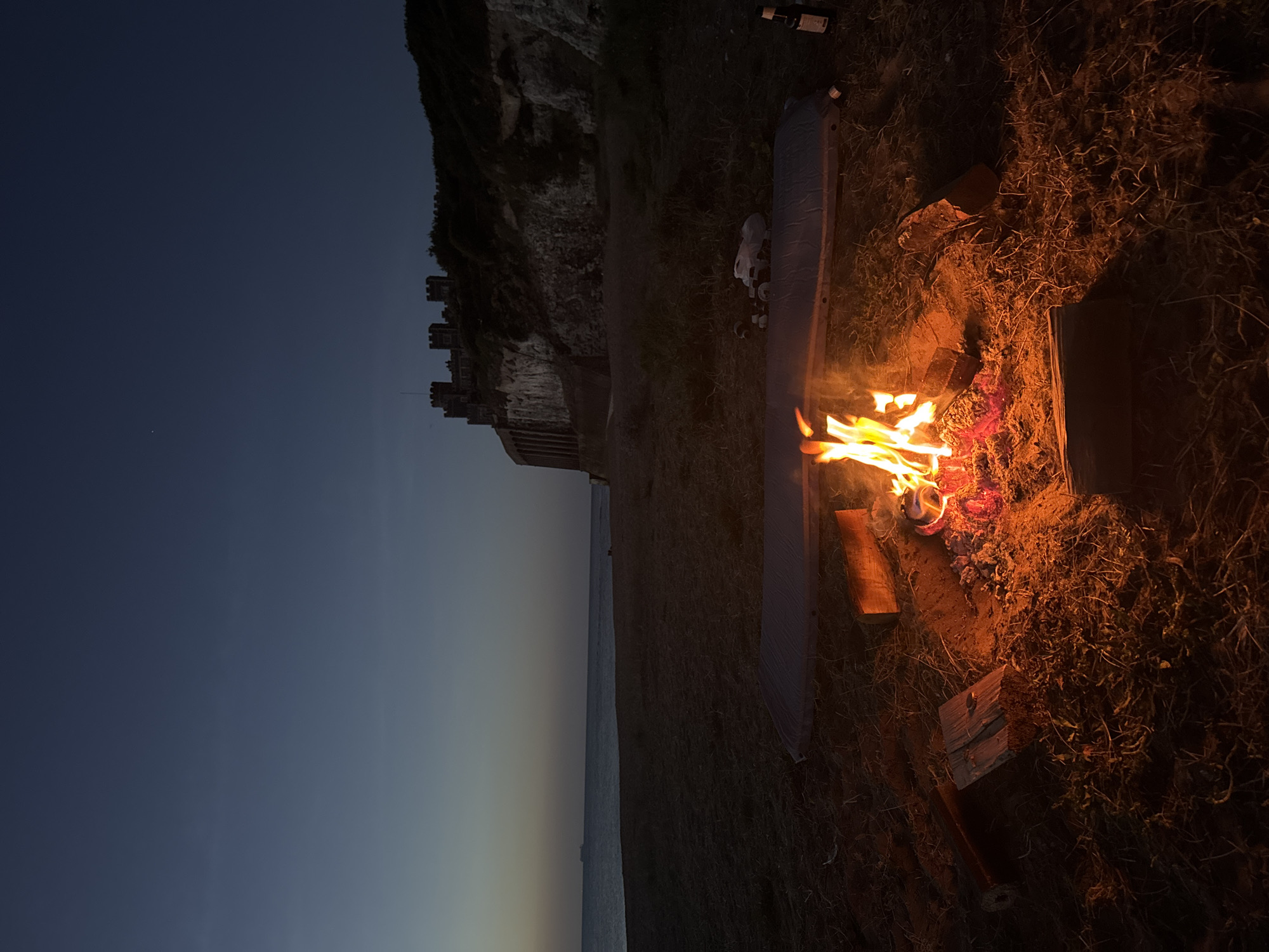 A campfire burns on a beach in Thanet, United Kingdom, at dusk. In the background, a castle sits atop a cliff overlooking the sea. The scene is peaceful and serene, with the flames of the fire casting a warm glow on the surrounding sand and debris. A camping mat and some bottles are placed nearby. A ship is visible on the horizon. The sky is a deep blue, with a few stars starting to appear.