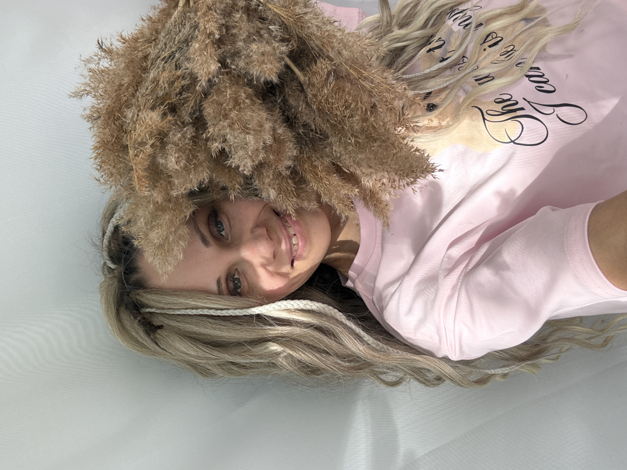 A young woman with fair skin and blonde hair with braided accents lies on a white surface, possibly a sheet or tablecloth. She is wearing a light pink long-sleeved shirt with text printed on it. A bundle of dried, brown ornamental grasses is positioned above her head. She is smiling. The location is not discernible as being in Rusanovo, Russia, based on the visual information in the image.