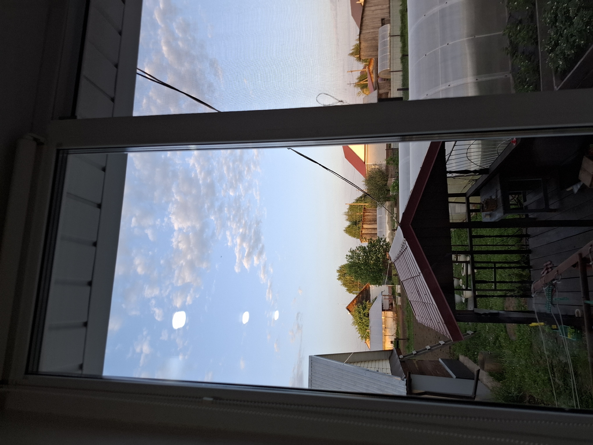 The image depicts a view from inside a building through a window or doorway frame. The exterior scene consists of houses, vegetation (trees and grass), and the sky with clouds. The buildings appear to be of residential or agricultural use, with one structure possibly a greenhouse. The sky is light blue with fluffy white clouds, suggesting a partly cloudy day. The presence of houses and greenery suggests a suburban or rural environment, potentially located in a residential area on the outskirts of Амзя, Russia. There are no people visible in the image.