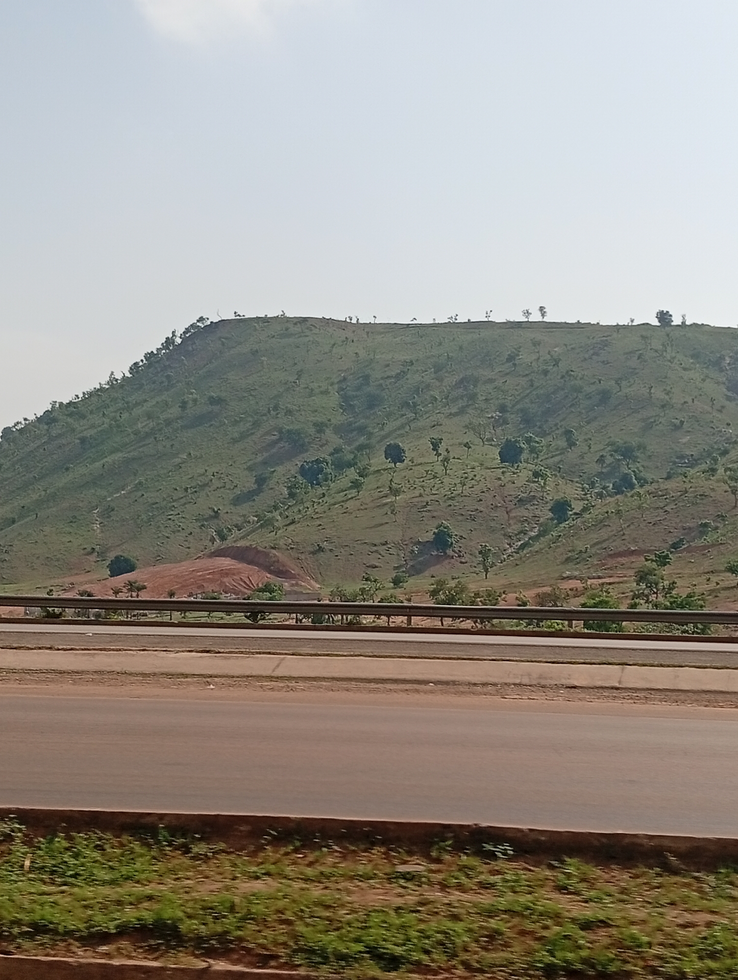 The image shows a hill with green vegetation and scattered trees, possibly in Kugbo, Nigeria, as indicated in the prompt. A section of exposed reddish-brown soil is visible on the hillside. A highway with a metal guardrail runs in the foreground. The sky is overcast with a light haze. A strip of green grass grows along the highway.