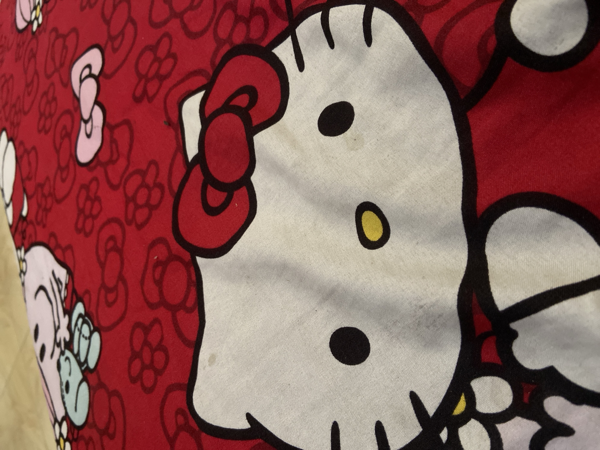 The image is a close-up, low-angle shot of a red fabric featuring a pattern of cartoon characters, primarily Hello Kitty and other characters in a similar style. The fabric appears to be a bedsheet or pillowcase. The main subject is a large depiction of Hello Kitty's face, rendered in white with black outlines, featuring a red bow on her head. Other smaller Hello Kitty faces and pink bows are visible in the background, interspersed with floral motifs. The fabric shows some signs of wear and staining, with visible spots of discoloration on the white areas of Hello Kitty's face. The lighting is somewhat dim, creating a slight shadow effect. There are no people or animals visible in the image. The image is taken indoors, likely on a bed or a surface where the fabric is laid flat. No specific location in Plosokandang, Indonesia, is depicted or implied by the image content.