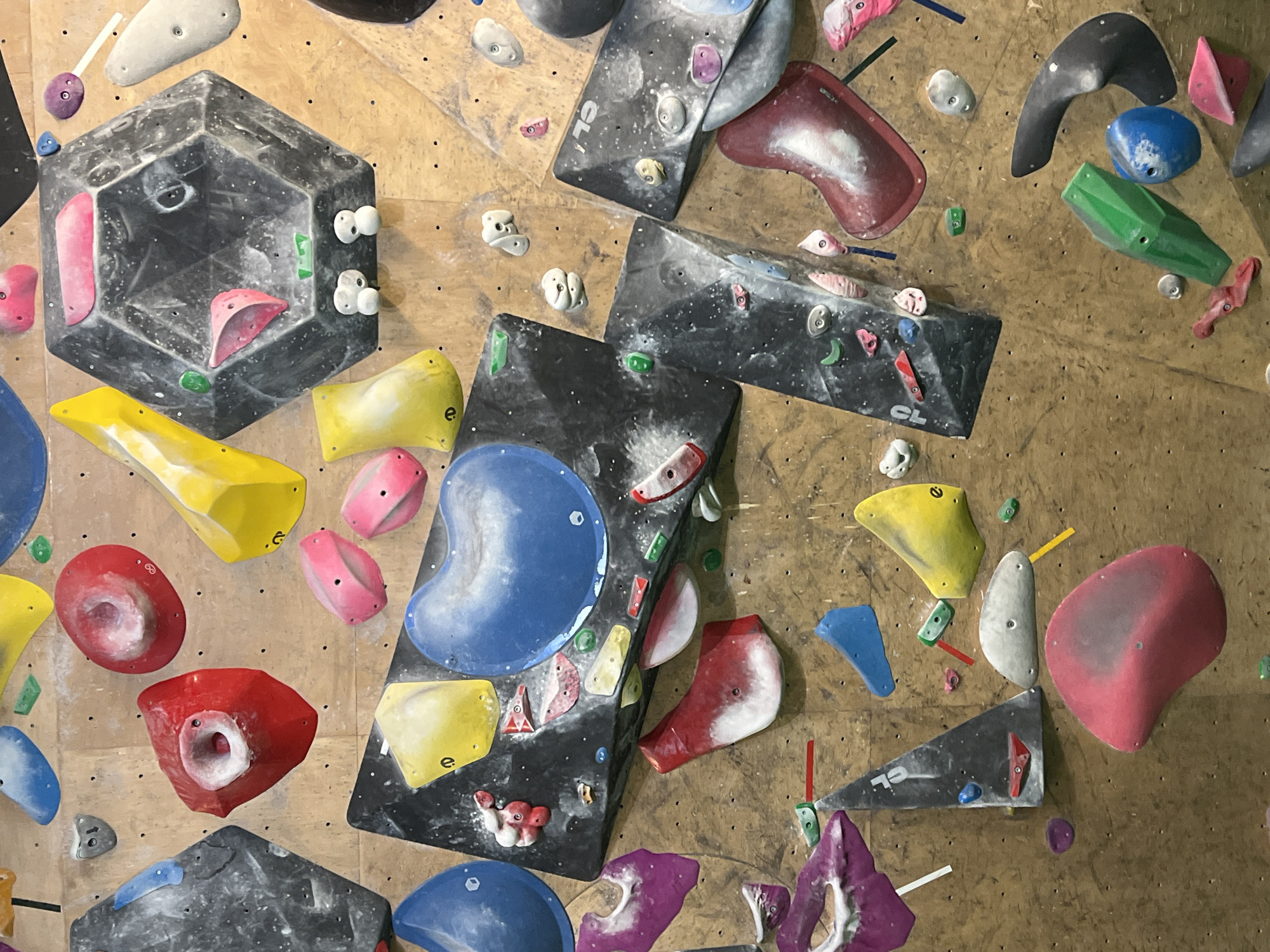 The image shows a climbing wall with various holds attached to it. The holds are of different shapes, sizes, and colors, including yellow, red, pink, blue, green, white, and grey. Some holds are solid colors, while others have patterns or textures. The wall itself appears to be made of wood, with a few small black dots visible. The city of Khimki, Russia, is not evident in the picture.