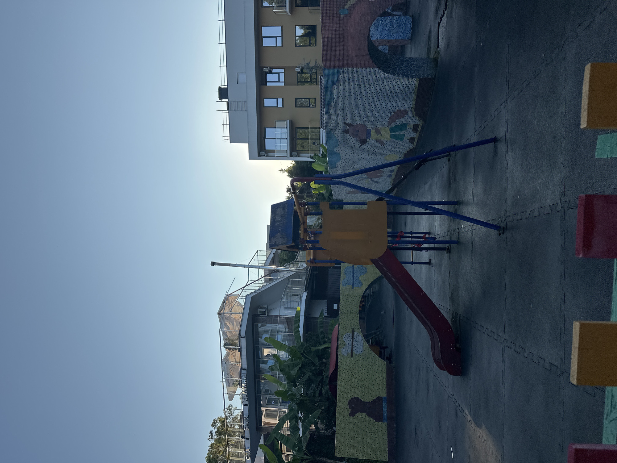 In Sochi, Russia, an outdoor playground is depicted under a clear blue sky. A red slide, part of a larger play structure with blue metal supports and a yellow platform, dominates the foreground. The ground is covered with dark grey interlocking rubber mats, showing some wetness. To the left, a yellow mosaic wall features a silhouette of a dog. To the right, a similar mosaic wall displays a cartoonish fox. Behind the playground, a multi-story building with a yellow facade and balconies, typical of Sochi's architecture, is visible. Further back, a more unusual building with a rounded, glass-enclosed upper level and exterior stairs is present, identified by text on its facade as "SPA БАНИ ХАММАМ." Greenery, including banana trees, surrounds the buildings. The scene appears to be set in the late afternoon, with the sun casting long shadows. There are no people visible in the image.