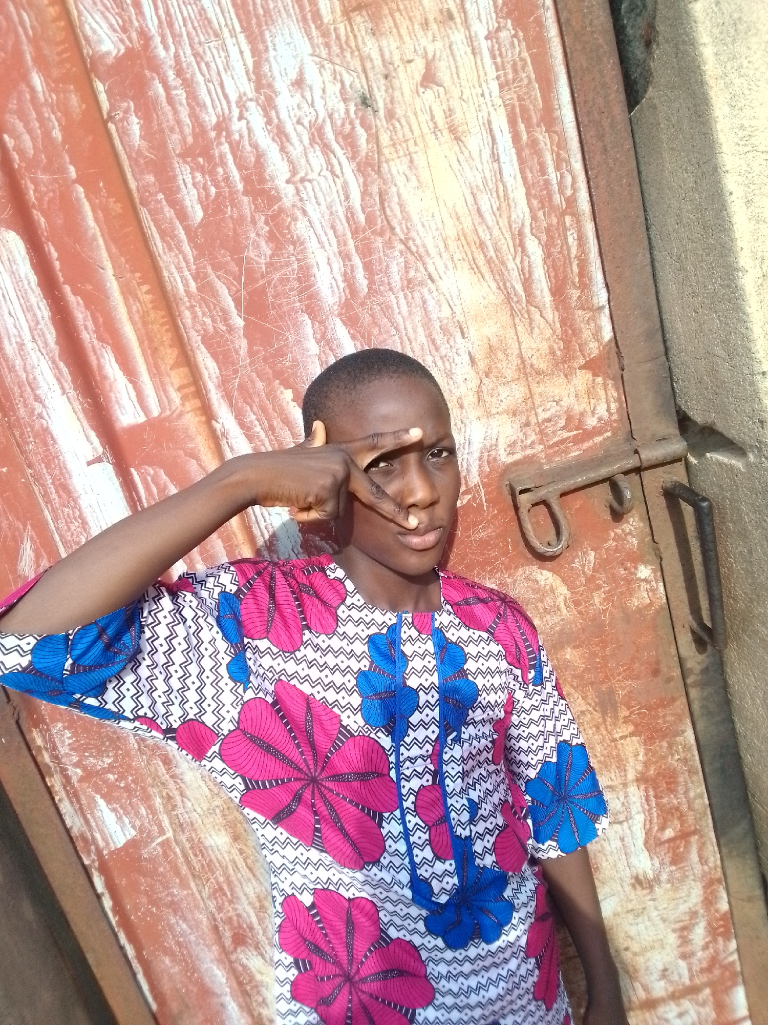 A young person stands in front of a textured, possibly metal, red door with a rusty latch and handle. The person is wearing a patterned shirt with pink and blue floral designs over a zigzagging white and black background. They are holding their right hand up, with their index and middle fingers extended to their right eye. The scene is brightly lit, casting shadows on the door and the adjacent wall. The location is estimated to be in the city of Orlu, Nigeria.