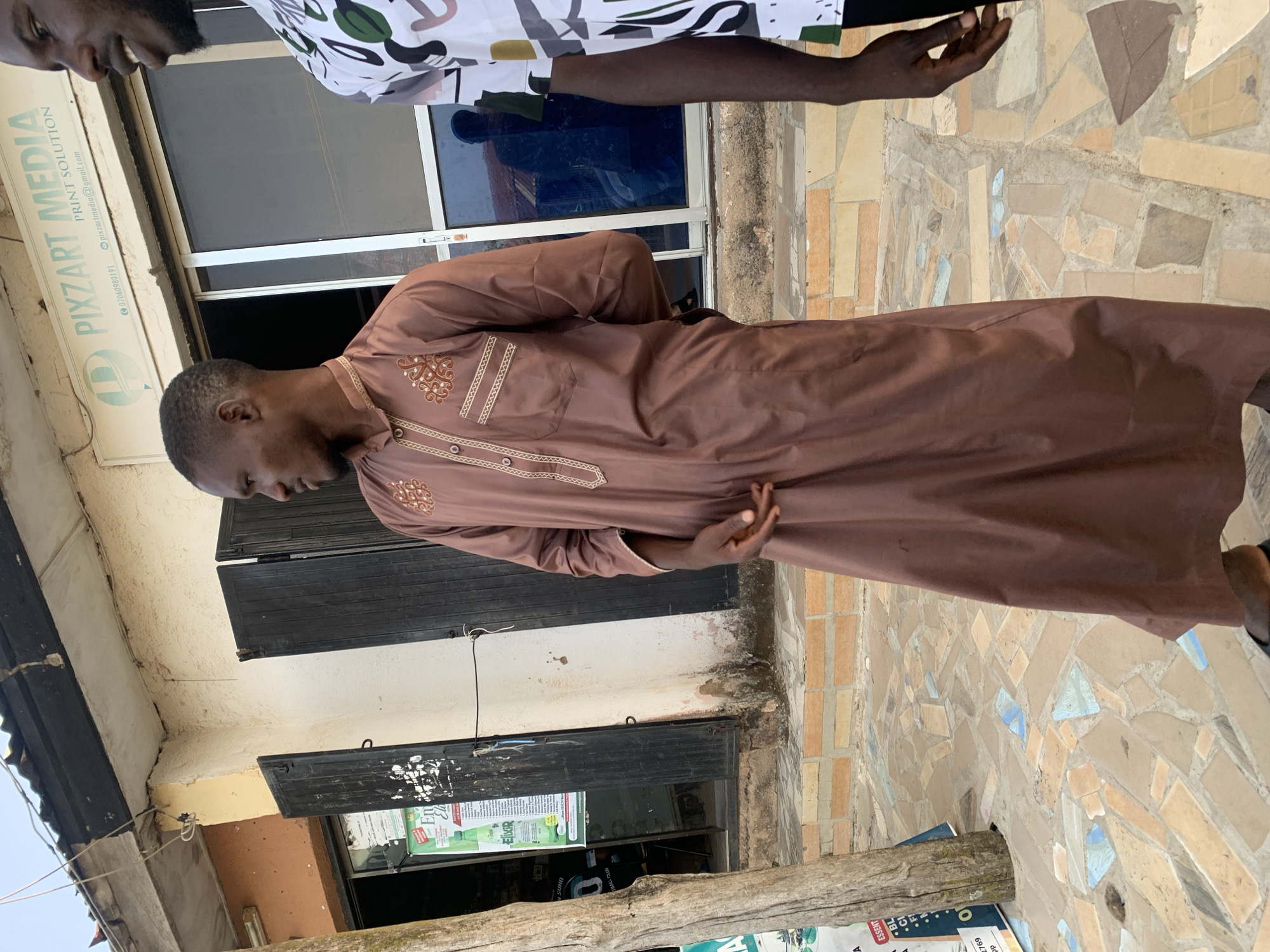 The image shows a man in a brown traditional robe standing outside a building in Jos, Nigeria. The building appears to be a print shop named "PIXZART MEDIA" with a sign displaying its contact information. Another man's head and arm, clad in a patterned shirt, are partially visible above the man in the robe. The sidewalk in front of the building is tiled with a mosaic pattern. The area seems to be a commercial street with posters and signs visible.