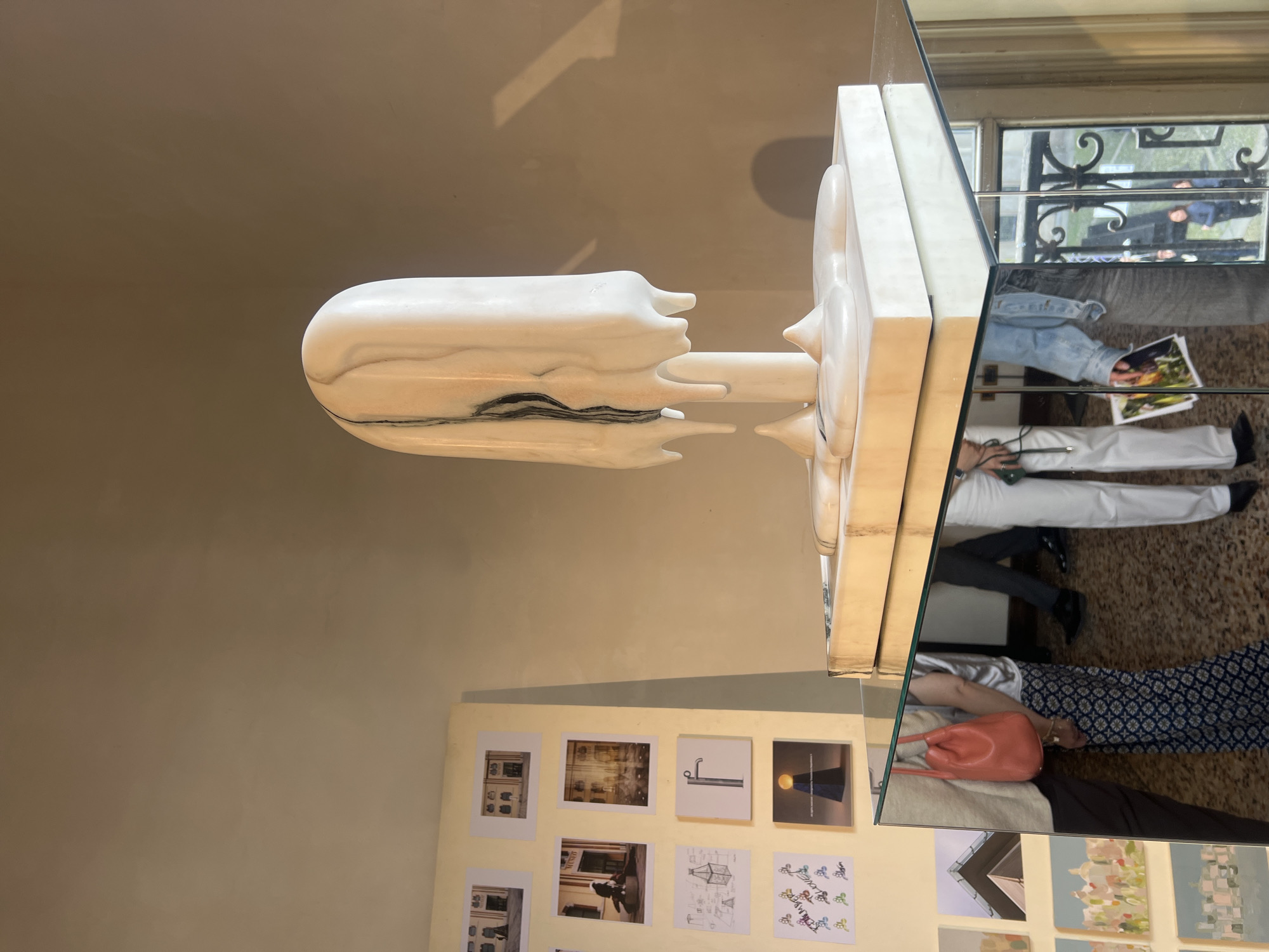 An indoor scene features a prominent white marble sculpture displayed on a reflective surface, possibly a mirrored tabletop or plinth. The sculpture, primarily white with striking black veining, especially near its base, has an organic, somewhat melting or dripping form. It consists of a tall, rounded rectangular upper section that narrows towards a slender central stem, which connects to a wider, flat base featuring rounded protuberances.
The reflective surface provides a fragmented view of the surroundings. Reflections of several people are visible: one person wears a light blue denim top and dark pants, holding a paper with an image; another is dressed in white pants and dark shoes; and a third wears a dark patterned skirt and a light top, carrying an orange/coral-colored handbag. These reflections suggest people are standing nearby, observing the display or moving through the space.
Beyond the immediate reflections of people, the mirrored surface also shows an outdoor scene through a window with dark, ornate grates, hinting at classical European architecture, consistent with a location like Milan, Italy. The lighting appears to be bright daylight.
In the background, a light yellow wall is adorned with an array of various artworks or photographs. These include images of interiors, architectural sketches (such as a lamp post or building outlines), and abstract or patterned illustrations. No discernible text is clearly legible on any of the visible items or artworks. The overall setting suggests an art exhibition, gallery, or a curated artistic space.