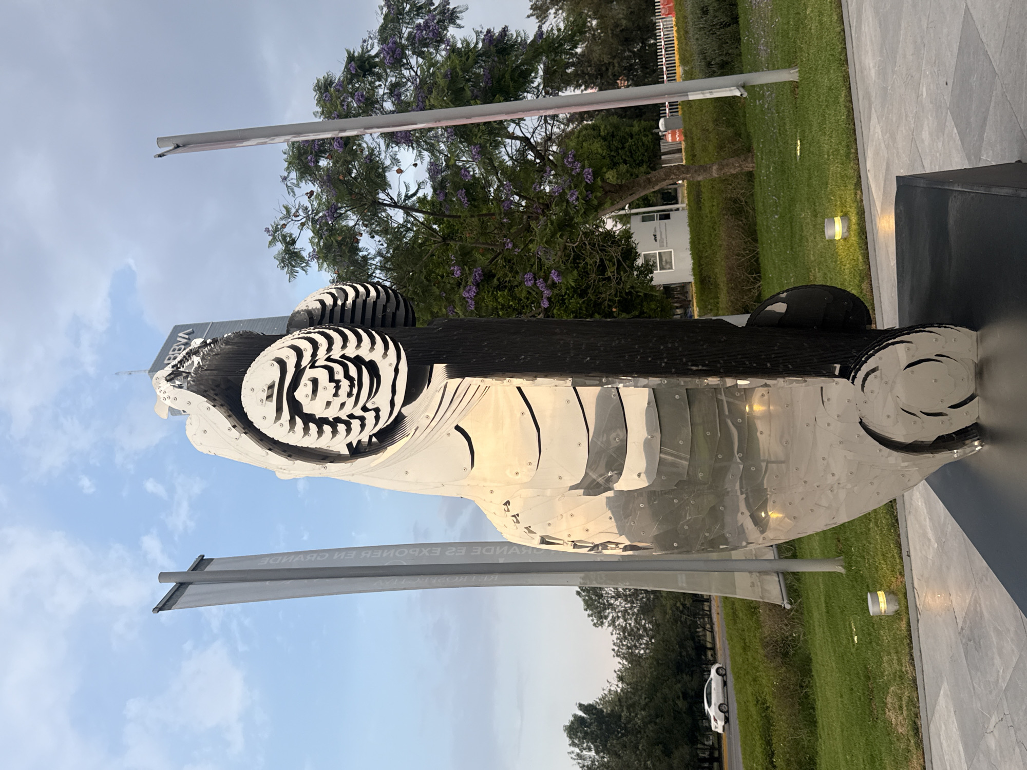 A photograph captures a modern, artistic sculpture that resembles a race car. The sculpture appears to be constructed from metallic materials, possibly stainless steel, with intricate cut-out designs that give the impression of movement. Its tires are black and white, with a striped pattern. The sculpture is located outdoors, possibly in a park or on the grounds of a building, as there is a well-manicured lawn in the background, along with trees and a white building that can be seen behind the sculpture. The sky is visible in the background. The sculpture is adjacent to a flag with some words on it, which is too blurry to be read. There are also outdoor light fixtures that are visible along the side of the sculpture's pedestal. The photograph was most likely taken in the city of San Bernardino Tlaxcalancingo, Mexico.