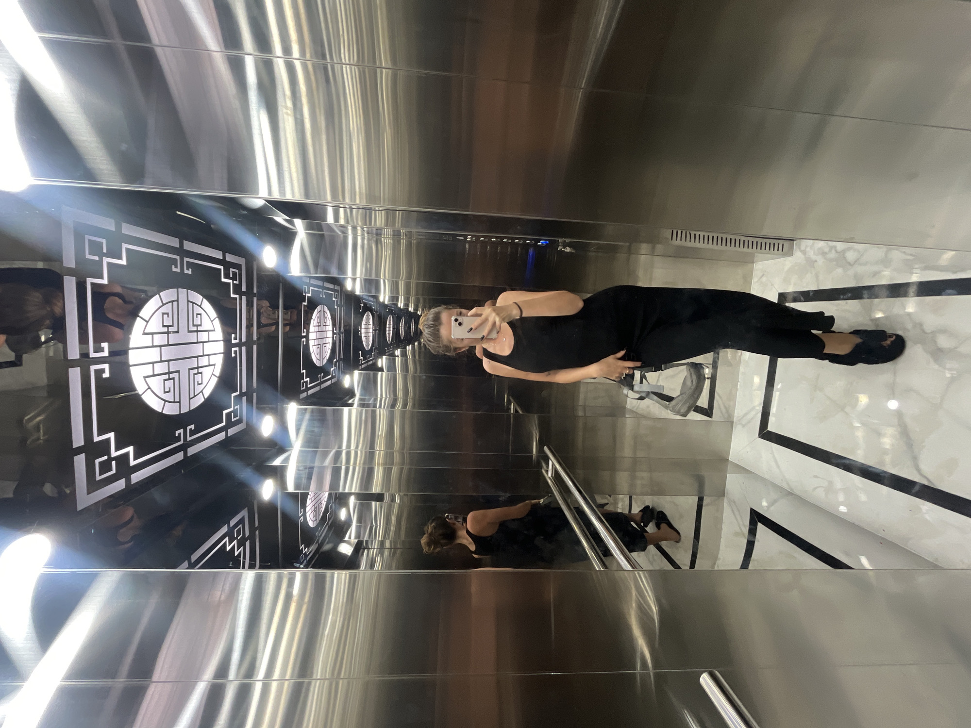 A full shot, captured from a low angle, shows a young Caucasian woman posing for a selfie inside a mirrored elevator. She has long brown hair pulled into a bun and is wearing a black tank top dress and black slip-on shoes. Her left hand is holding her phone, while her right hand rests on her hip. The elevator's walls are made of brushed stainless steel, and the ceiling features a black background with intricate white geometric patterns and circular illuminated designs. The floor is white marble with black borders. In the background, her reflection is visible in the mirrored walls, including a glimpse of a woman in a similar black dress standing by the elevator doors on the left. The elevator appears to be located in a modern building, likely a hotel or residential complex, possibly within the city of Nha Trang, Vietnam, based on the provided context about Bac Nha Trang Ward. The lighting is bright and reflective, creating a somewhat disorienting effect due to the mirrored surfaces.