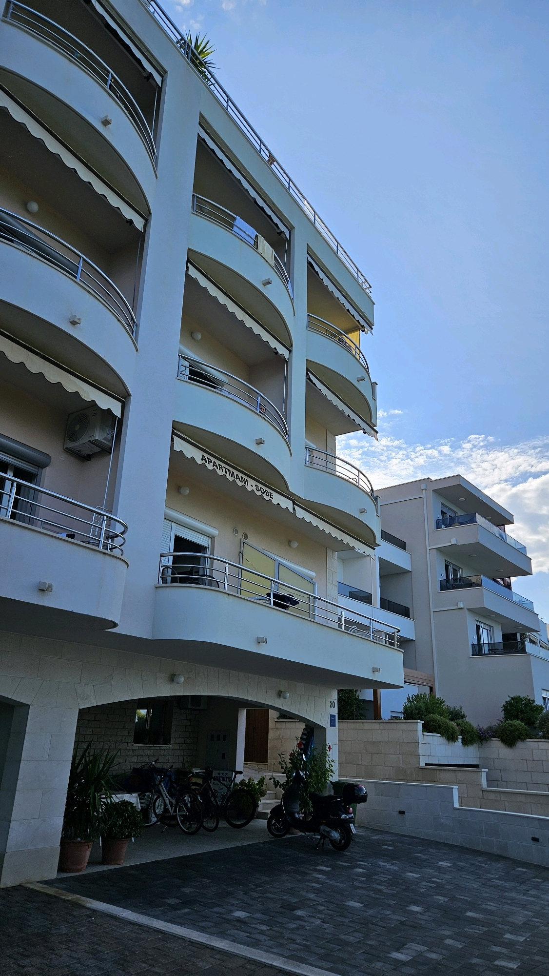 The image depicts the exterior of an apartment building in Općina Tučepi, Croatia. The building has multiple levels with curved balconies, each equipped with retractable awnings and metal railings. The facade is painted a light color, and an air conditioning unit is visible on one of the balconies. A sign above an entrance reads "APARTMANI - SOBE." In the foreground, a paved area serves as a driveway or parking space. A black scooter is parked on the right side, and a group of bicycles are situated under a covered entryway on the left. Several potted plants are placed near the entrance. Another building, similar in style but with a more angular design, is visible to the right, also featuring balconies and modern architecture. The sky above is blue with scattered white clouds. The scene conveys a sense of residential architecture in a coastal or Mediterranean setting.