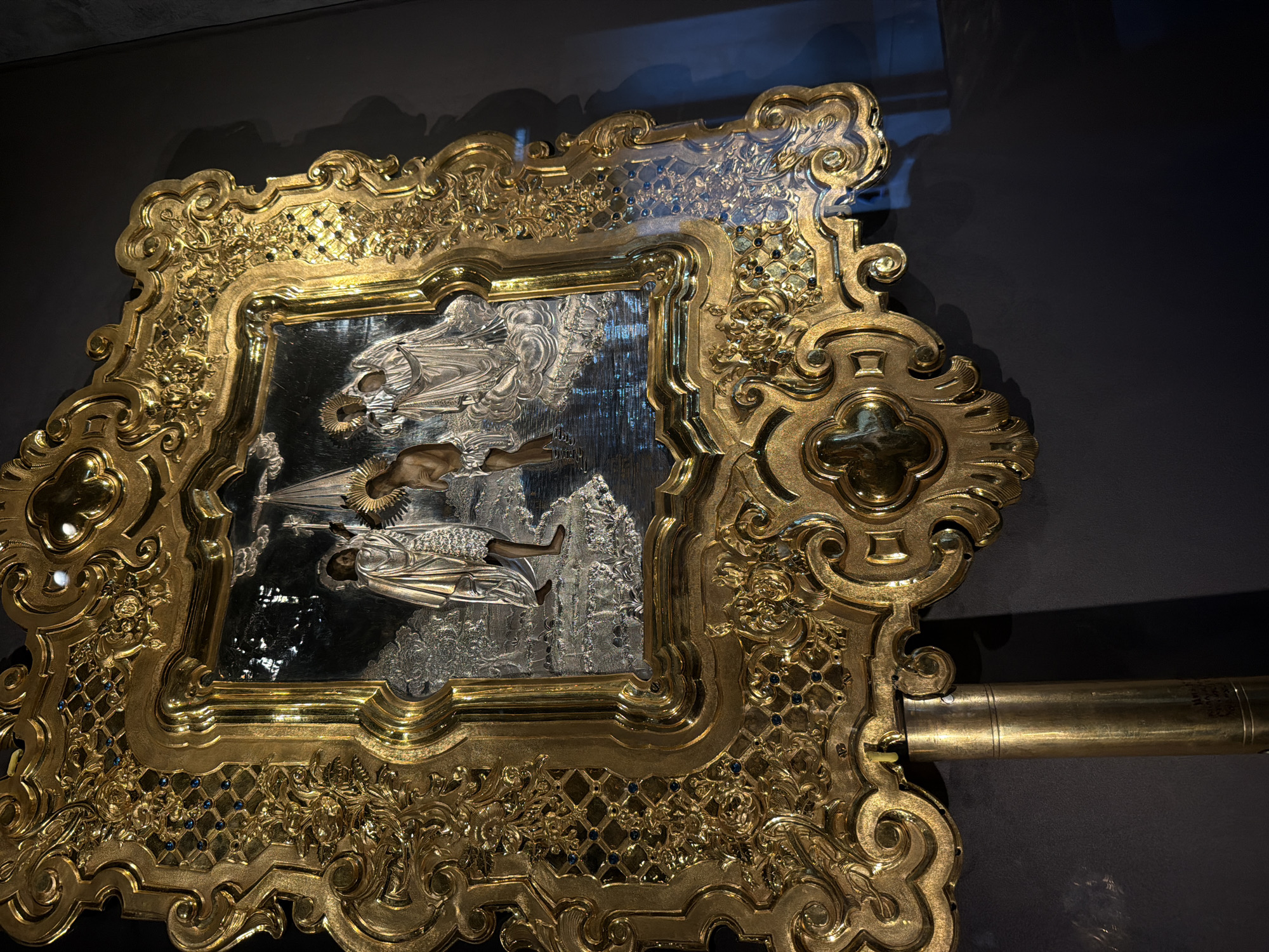 A gold-framed icon is displayed against a dark gray background. The icon itself depicts figures in what appears to be a landscape. The frame is ornate, with a lattice pattern and decorative flourishes. To the right of the frame is a gold-colored cylindrical object, possibly a support or handle. No people or actions are directly visible in the image. The image was taken inside a museum in the city of Perm, Russia.