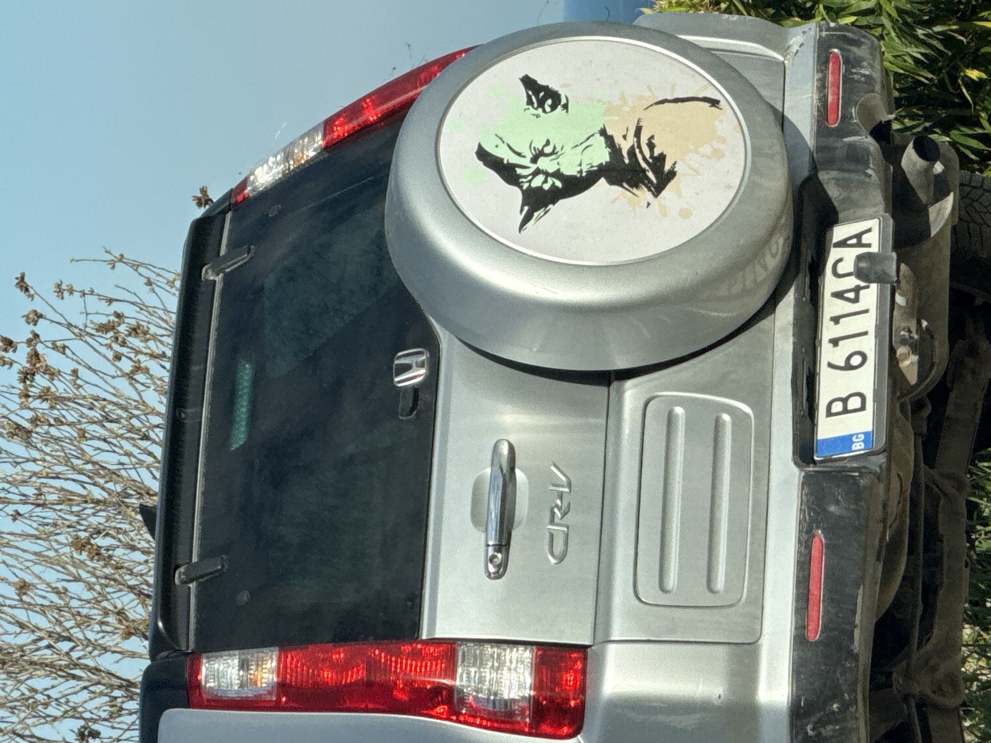 The image shows the rear of a silver Honda CRV. A spare tire cover on the back features a stylized image of Yoda from Star Wars. The license plate reads "B 6114 CA" with a "BG" indicator. The car's location is assumed to be in Varna, Bulgaria. The environment is difficult to ascertain, with only a few sparse branches visible, suggesting an outdoor location.