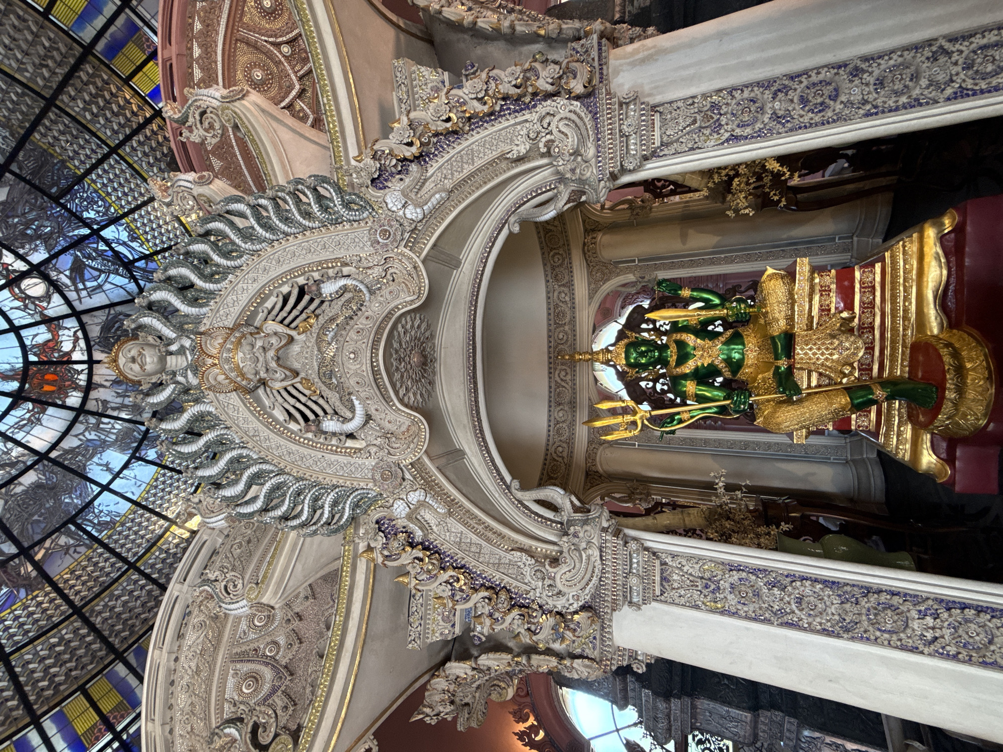 The image depicts an ornate interior, potentially a temple or religious site in Samrong Nuea Subdistrict Municipality, Thailand. A green and gold statue stands within a decorated alcove. Elaborate carvings and mosaics adorn the walls and ceiling, featuring intricate patterns and designs. A stained-glass dome-like structure appears in the upper left portion of the frame. The statue is a figure in either gold or bright green color, and is sitting on an elevated platform also in gold. It is inside what appears to be an archway and a room that is ornately designed with carvings.