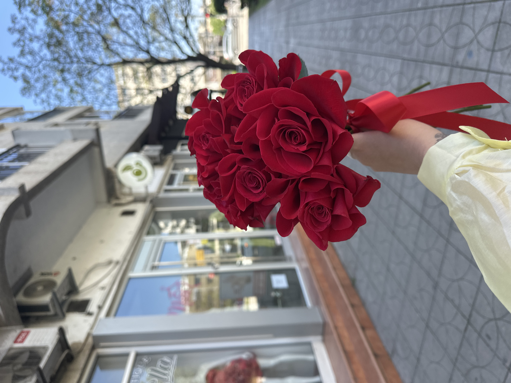 A bouquet of red roses, tied with a red ribbon, is held in a person's hand against a blurred urban backdrop. The person is wearing a light-yellow shirt or sleeve. The roses are prominently displayed, and the hand is positioned to the right of the frame. Behind the roses is a building with glass windows. A tree branch is visible in the upper left. The location is Šušanj, Montenegro. A patterned walkway is visible in the foreground.