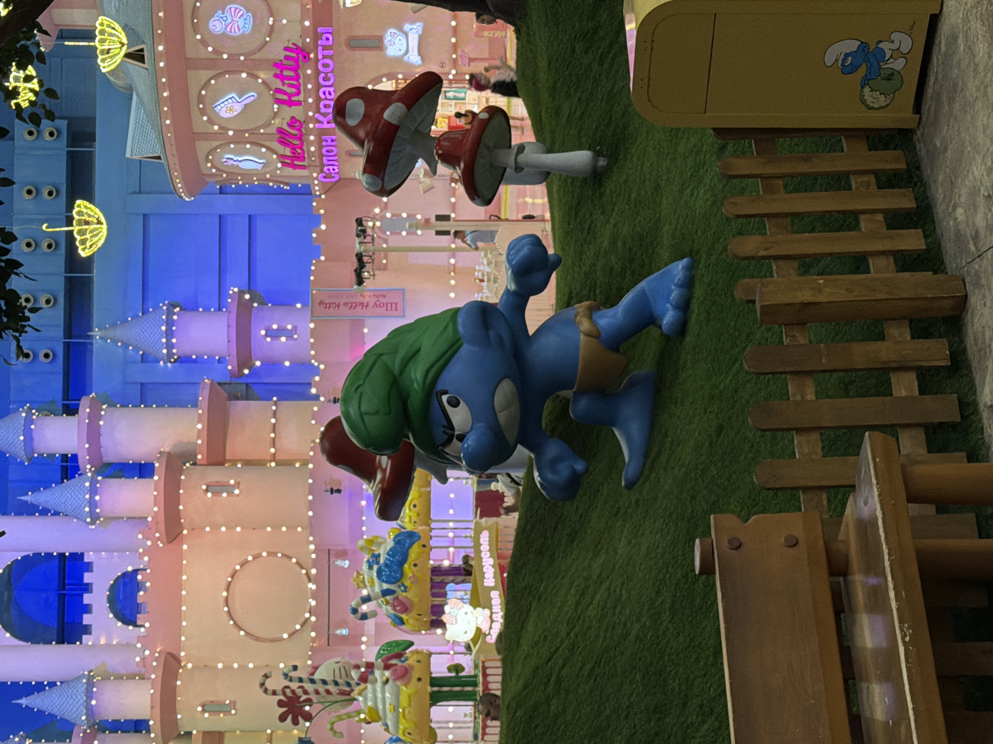 This image depicts an indoor amusement or entertainment area, likely a theme park or mall attraction, with a cartoonish, brightly lit aesthetic. In the foreground, a large, angry-looking blue Smurf figurine is positioned on a green, artificial turf-covered slope. Behind the Smurf, several red and white mushroom-shaped props, also appearing to be decorative elements, are visible. Further in the background, a pink castle-like structure adorned with numerous small white lights is prominent. To the right of the castle, a circular pink building with illuminated signs that read "Hello Kitty" and "Салон Красоты" (Beauty Salon) can be seen. Various other decorative elements, including candy-cane themed props and figures that resemble Hello Kitty, are scattered throughout the scene. The overall environment is artificial and designed for visual appeal. There is a wooden bench and a small wooden fence in the lower part of the frame, and a yellow trash bin with a Smurf decal on the right side. Several people are visible in the background, appearing to be visitors or staff, but they are small and indistinct. The lighting is vibrant, with significant use of pink, blue, and yellow hues. The specific location is identified as Vidnoye, Russia.