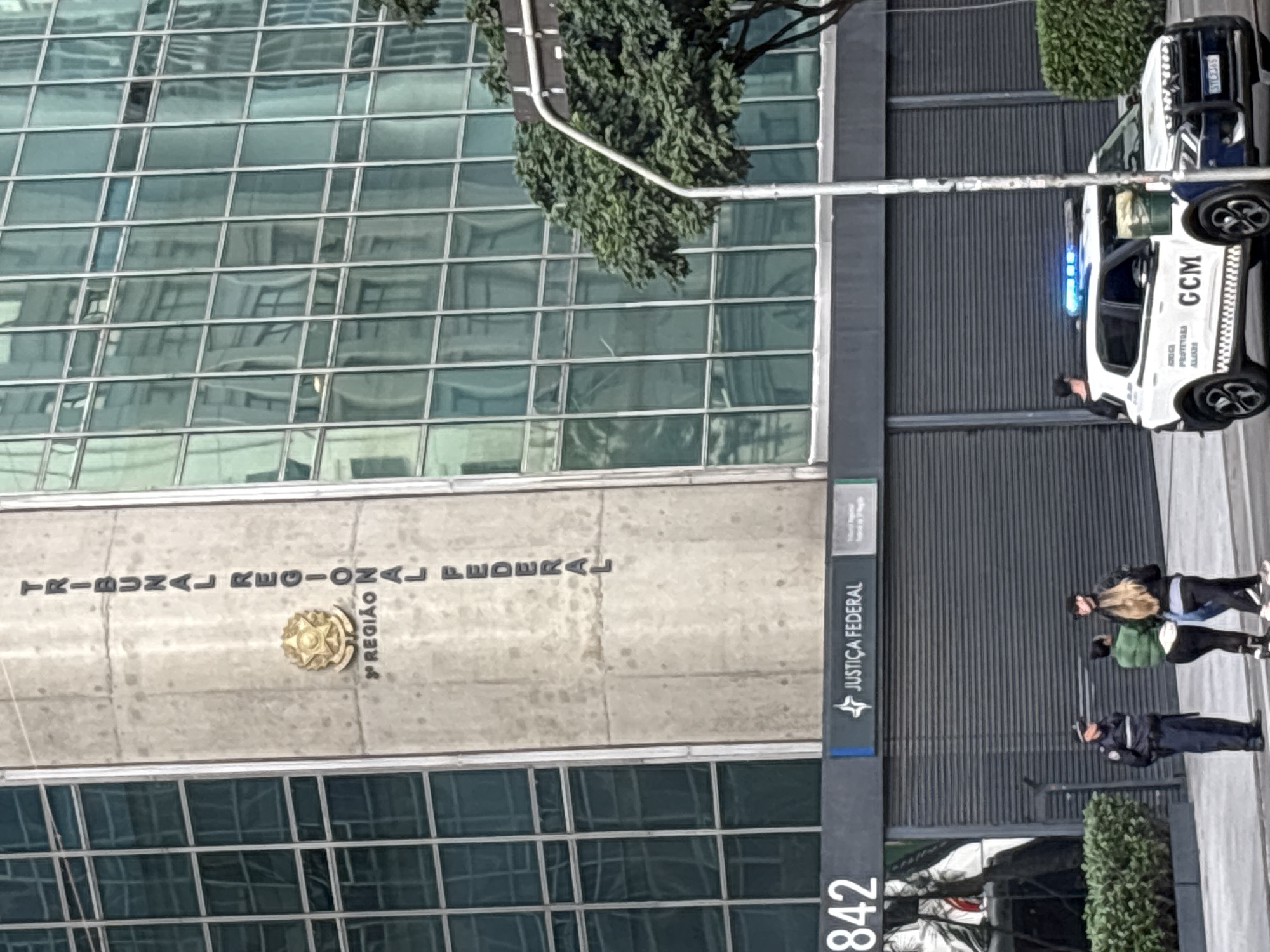The image shows the exterior of a building with a prominent sign reading "JUSTIÇA FEDERAL" and "TRIBUNAL REGIONAL FEDERAL 3ª REGIÃO". A police vehicle with flashing blue lights, marked with "GCM", is parked in front of the building, suggesting a police presence. Three individuals are visible near the building entrance, one in a police uniform and two civilians. The building is modern with a glass facade and a concrete section displaying the federal seal and the text. The scene appears to be a public street in São Paulo, Brazil, indicated by the architectural style and the Brazilian police vehicle.