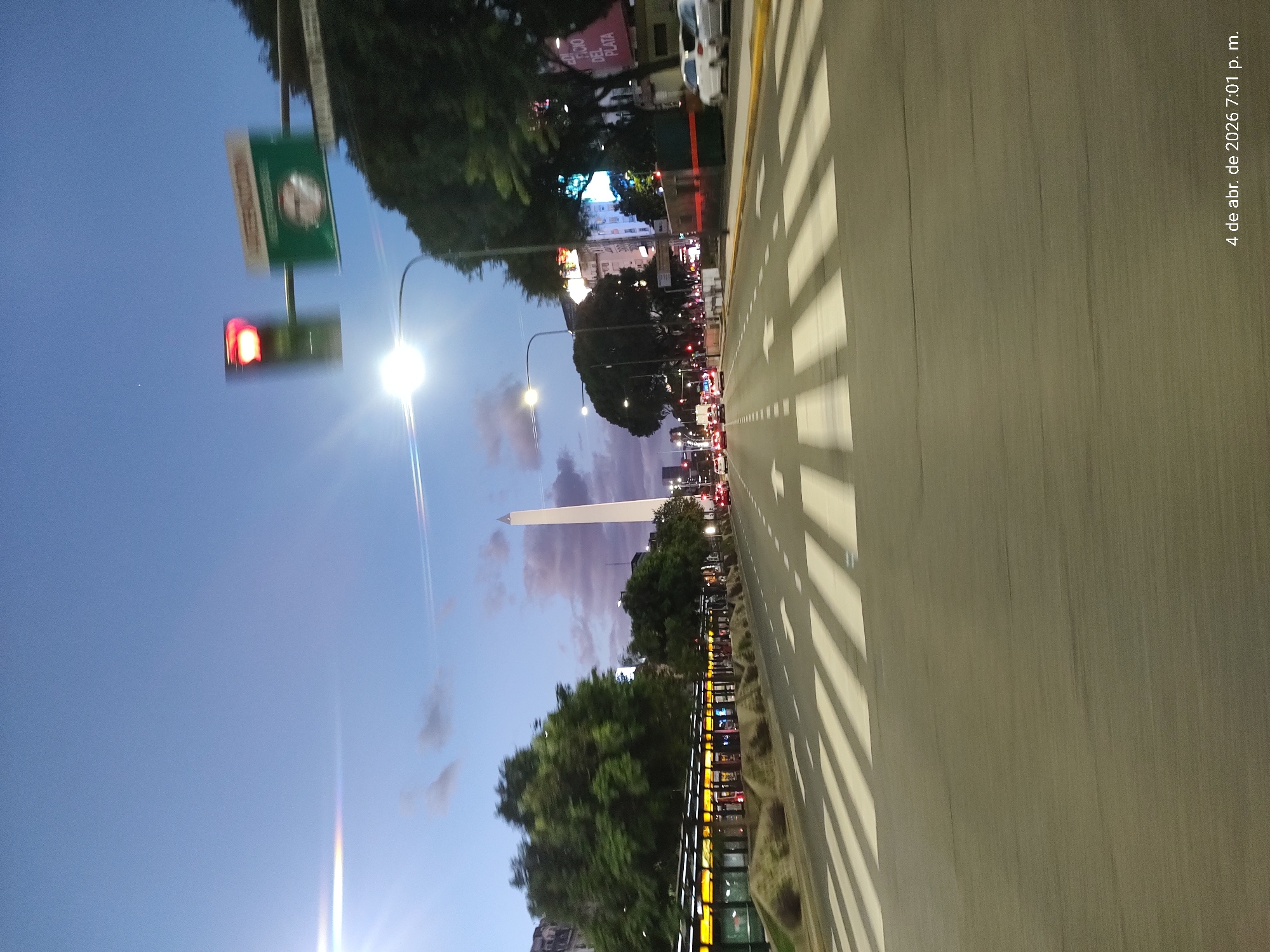 The image captures an urban scene at dusk in Buenos Aires, Argentina. The Obelisco, a prominent city landmark, stands tall in the distance. The view is from a street-level perspective, showcasing a multi-lane road with zebra crossings, illuminated by streetlights. Tree shadows create linear patterns on the road surface. A sign indicating "Obelisco" is visible. Traffic signals are present, with one displaying a red light. There are trees lining the avenue, and buildings are illuminated in the background. The date and time "4 de abr. de 2026 7:01 p. m." is visible on the right side of the image. There is traffic on the road.