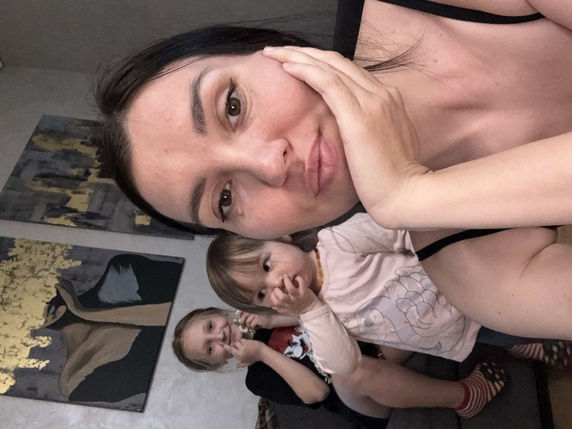 A selfie-style photograph captures a woman with dark hair and fair skin, holding her face with one hand. She is positioned at the top of the frame, slightly angled. Beneath her, two young children are visible. One child, wearing a pink long-sleeved shirt, has fingers near their mouth. The other, in a black shirt, appears to be holding something small near their face. The setting includes a wall adorned with artwork in the background. Although there is no way to determine the specific location as Koltushi, Russia based on the image alone, this is the specified location.