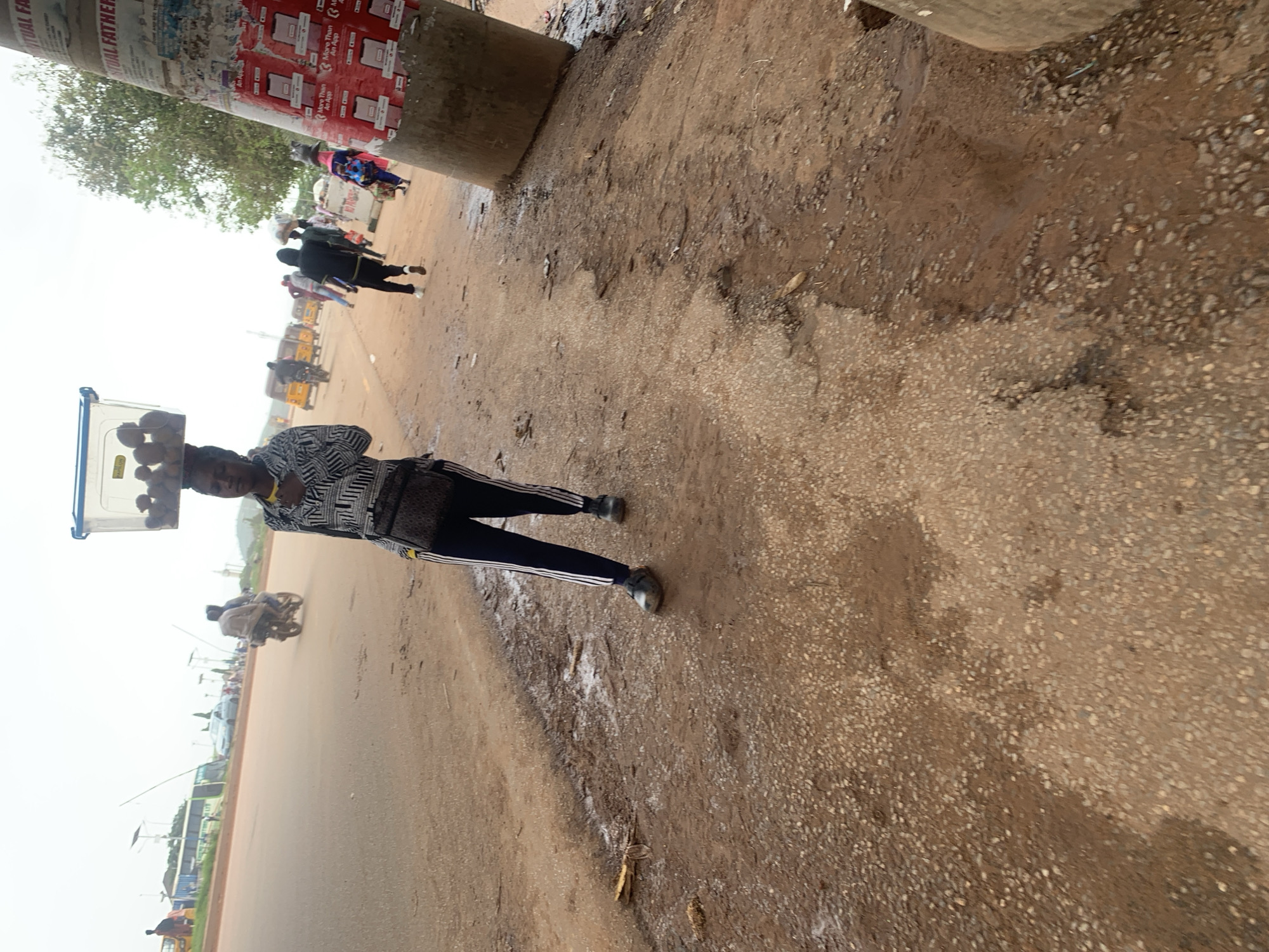 A full shot captures a young woman walking on a dirt roadside in Nyanya, Nigeria, carrying a plastic tub filled with fruits on her head. She is wearing a black and white striped shirt, dark pants with white stripes on the sides, and black shoes. A dark crossbody bag is slung over her left shoulder. In the background, vehicles including a bus, cars, and motorcycles are visible on a paved road. Pedestrians are also seen walking and carrying loads. A utility pole plastered with posters stands to the right of the frame. The ground is a mix of dirt and gravel with some puddles. The sky is overcast.