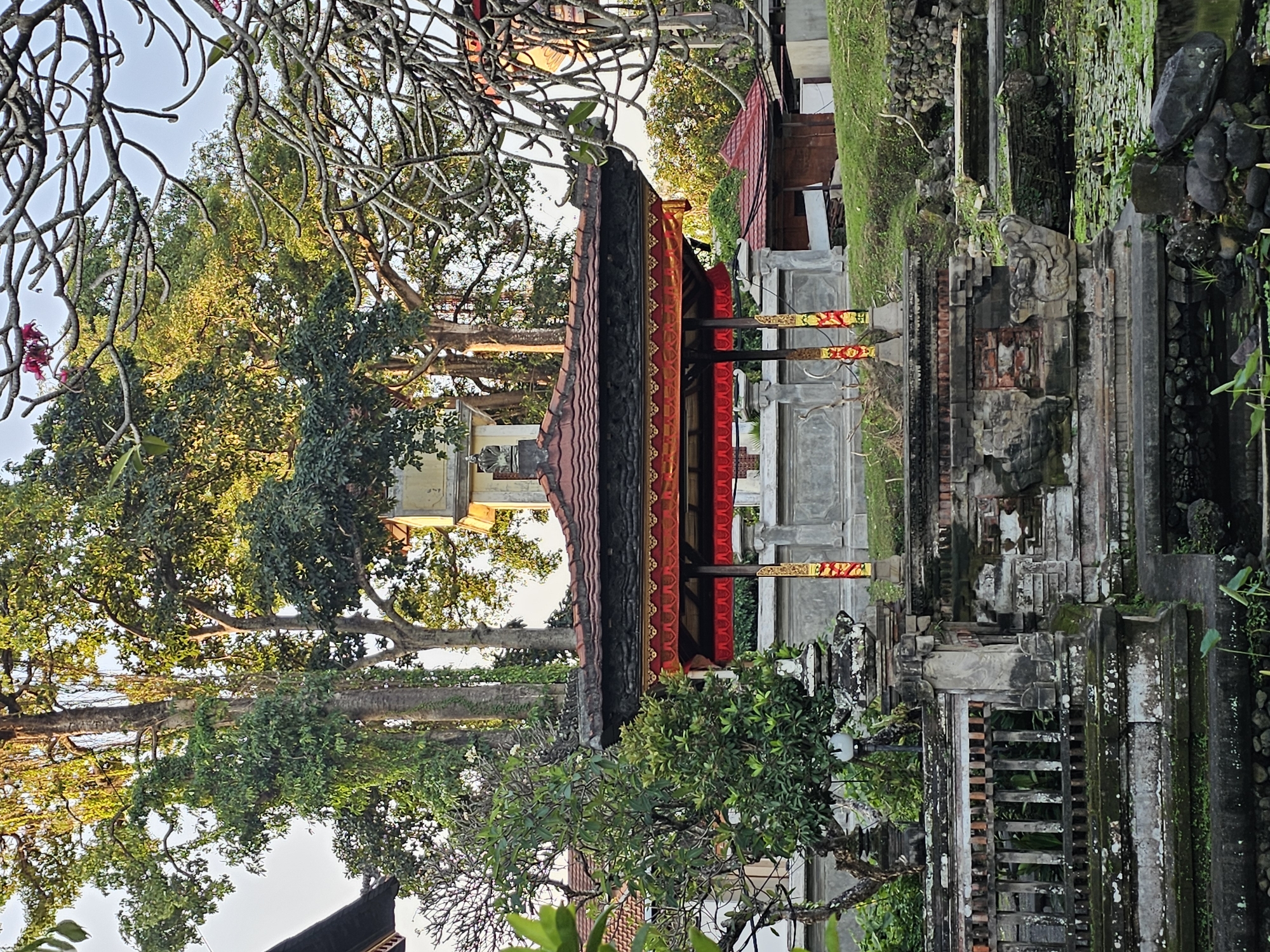 The image depicts an ornate pavilion with a red and gold tiled roof, supported by decorative pillars. The pavilion is set within a lush, green environment, likely a garden or temple complex. Large trees with dense foliage dominate the background, their branches extending into the clear sky. In the foreground, there's a stone structure with weathered carvings and a decorative railing, suggesting an ancient or traditional architectural style. A small pond or water feature with lily pads is visible at the bottom, further enhancing the serene atmosphere. The overall impression is one of tranquility and cultural richness. This scene is likely from Denpasar, Indonesia, a city known for its rich cultural heritage and numerous temples and traditional buildings.