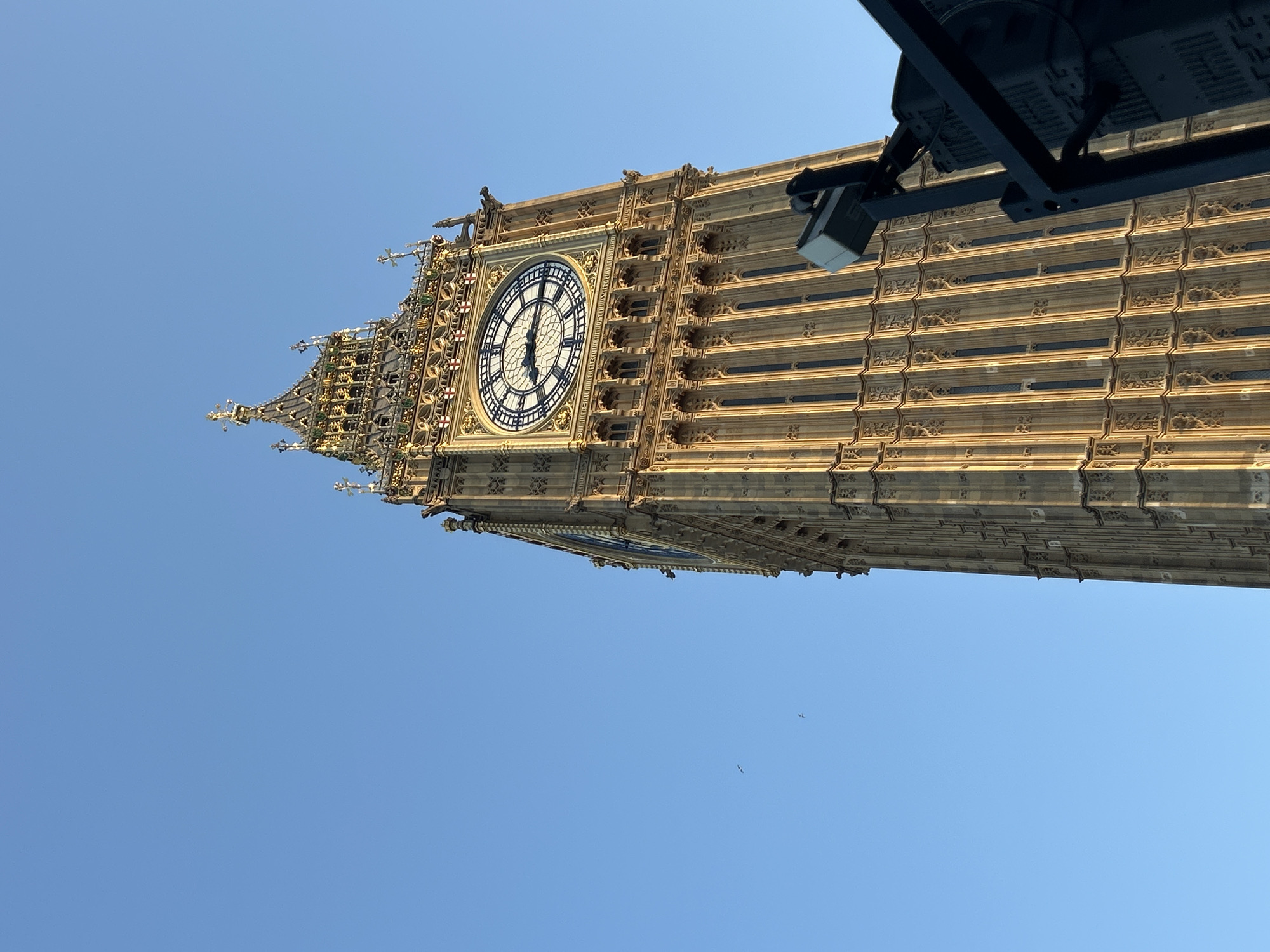 The image depicts the Elizabeth Tower, commonly known as Big Ben, in London, United Kingdom. The tower is viewed from a low angle, showcasing its intricate architectural details against a clear blue sky. The clock face is prominently visible, indicating a time around 6:03. Some modern lighting or camera equipment is mounted on the side. There are a few birds flying in the background, in the lower half of the image. The lighting suggests a sunny day.