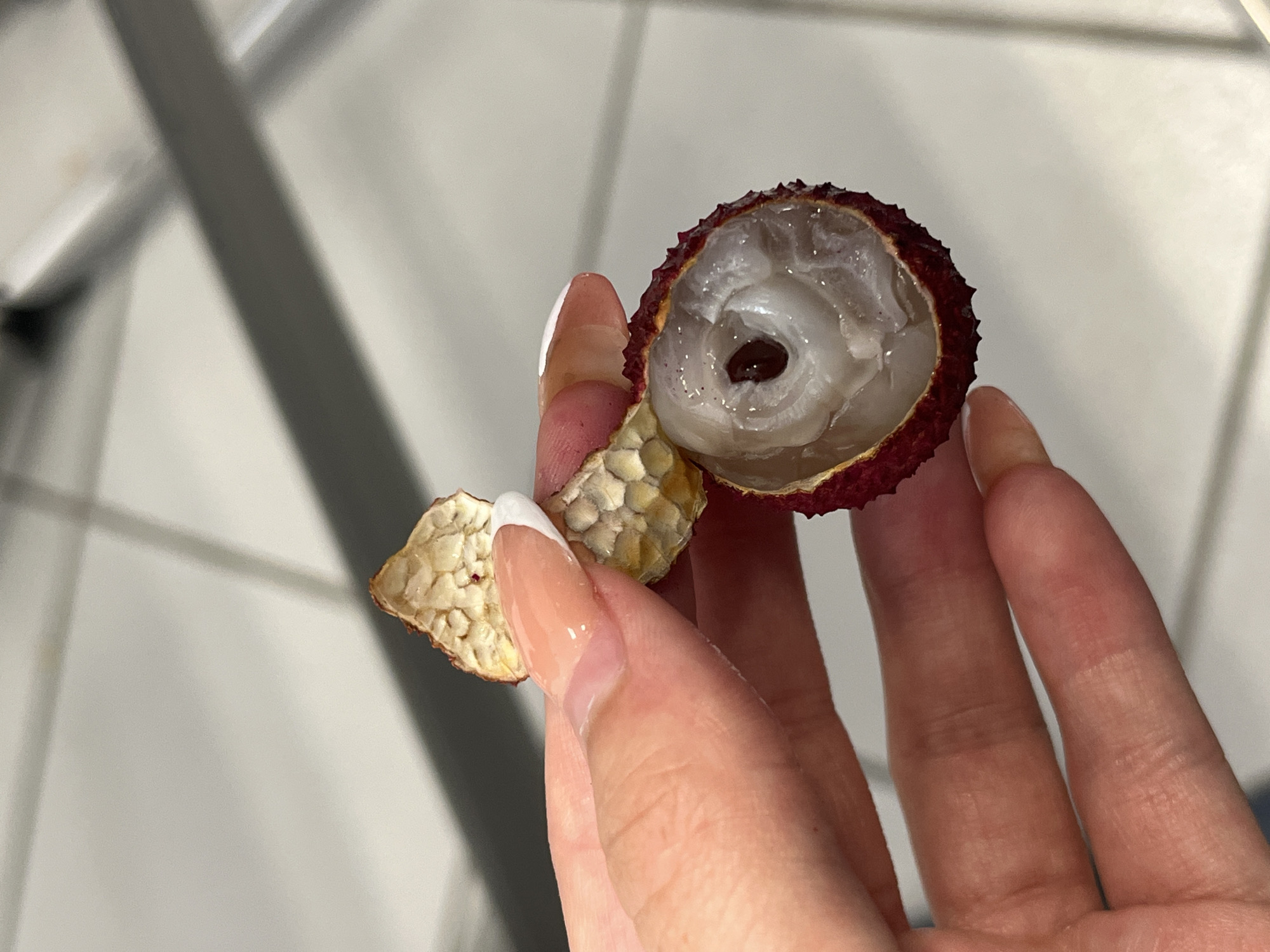 A hand holds a partially peeled lychee fruit. The lychee is a translucent white with a dark brown seed visible inside. The peel is a mottled tan and beige. The background is a blurred white and gray. The location is not discernible from the image.