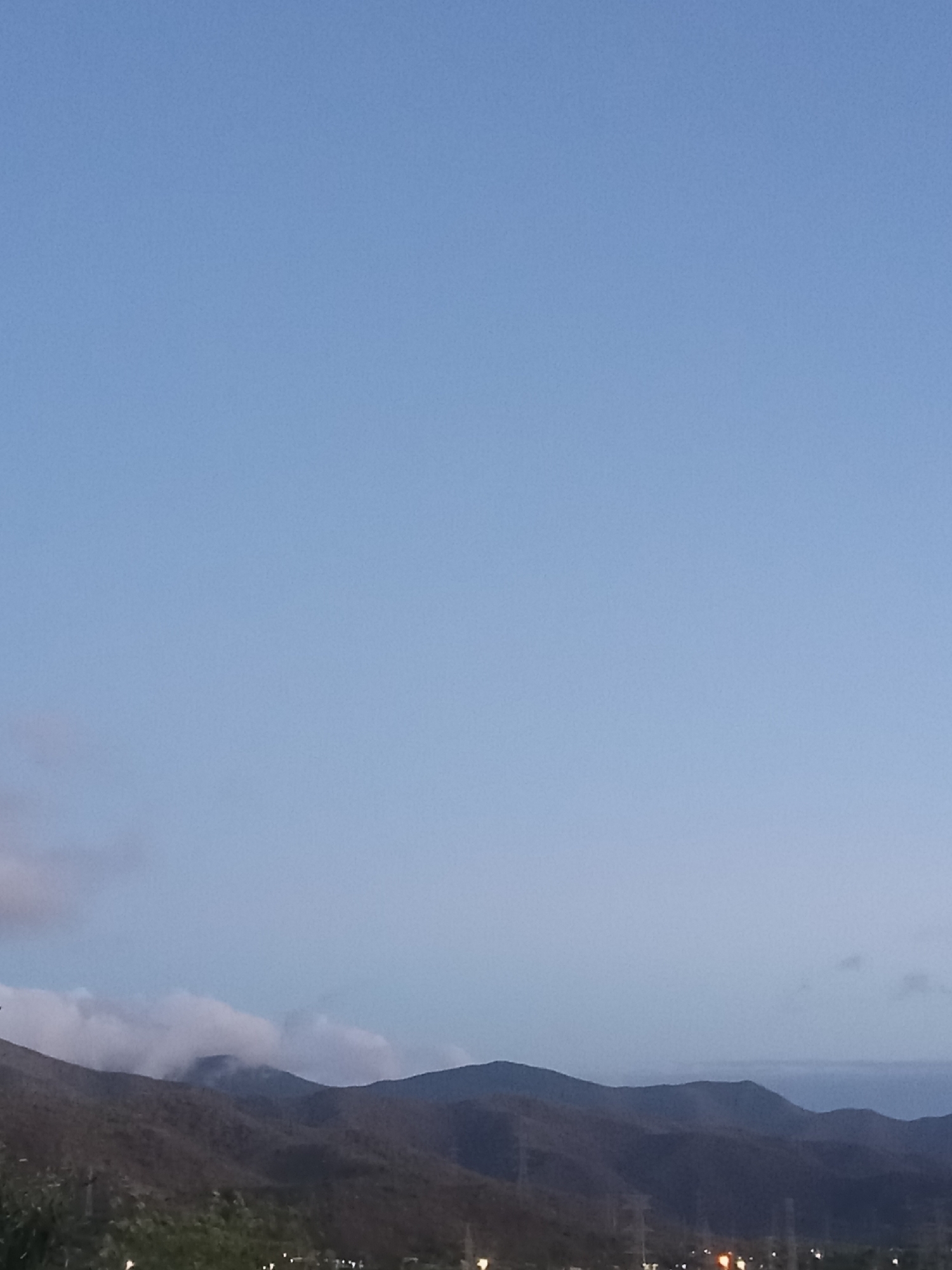 The image captures a twilight or dusk scene in a hilly or mountainous region, potentially near Altagracia, Venezuela. The sky occupies the majority of the frame, exhibiting a gradient from a lighter blue towards the top to a slightly darker hue closer to the horizon. The horizon is defined by a series of overlapping mountain ridges, which appear dark and silhouetted against the fading light. Some low-lying clouds are visible, partially obscuring the peaks of some of the mountains. In the foreground, a dark, indistinct mass suggests vegetation and possibly urban infrastructure, including faint lights that might be from streetlights or buildings. The overall impression is one of a peaceful, natural landscape transitioning into night.