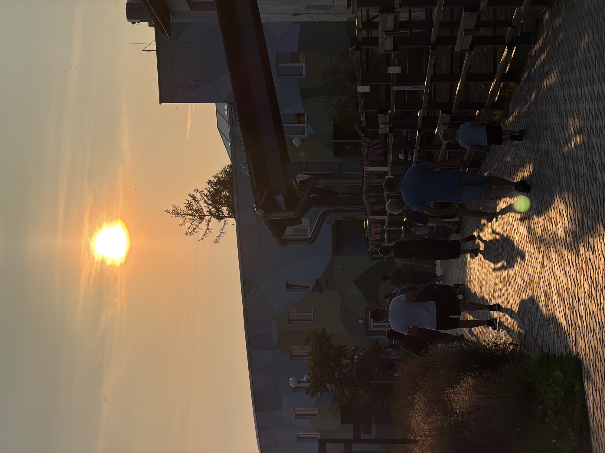 This media file captures a sunset scene in Perm, Russia, with a group of people walking along a paved walkway. The sun, a bright orb with rays extending outwards, is visible in the hazy, orange sky, indicating it is either sunrise or sunset. Long shadows are cast by the people and a wooden structure with horizontal slats, suggesting the sun is low in the sky.
The subjects are a group of individuals, some of whom are walking in the same direction along the walkway. Their attire is casual, consisting of t-shirts and shorts, indicating warm weather. The building in the background has a modern, somewhat abstract design with a color-blocked facade featuring various shades of blue, green, and grey. A small tree with wispy branches is silhouetted against the sky. The walkway is paved with brick-like stones, and there is lush greenery bordering the path.
The overall mood is peaceful and relaxed, possibly suggesting an evening stroll or a gathering after a day's activities. The low sun and hazy atmosphere create a warm, golden light that bathes the scene. No visible text is present in the image.