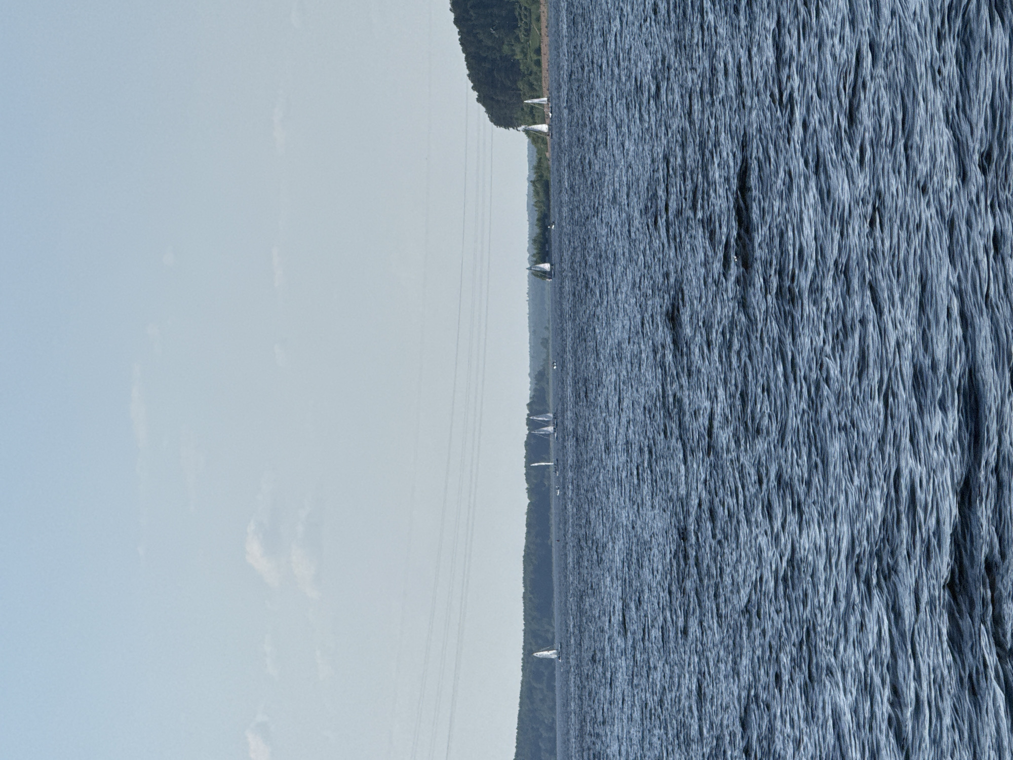 The image shows a body of water, likely a lake or reservoir, adjacent to a green shoreline. Several small sailboats are visible scattered across the water. On the far left of the image is a clear sky with sparse clouds. The water surface is textured with small waves. The location is potentially the Izhevsk Reservoir in Izhevsk, Russia.