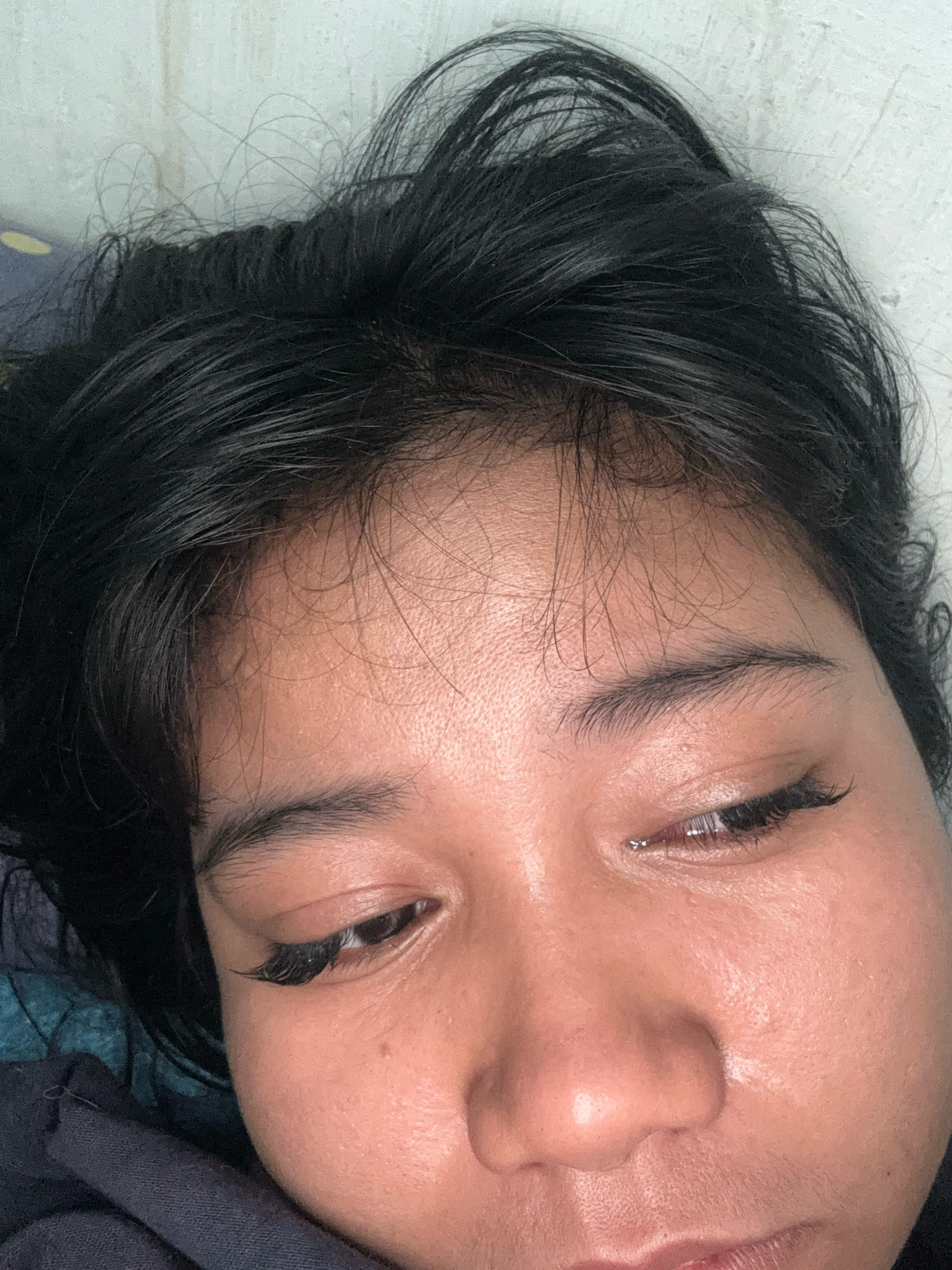 This is a close-up, eye-level, indoor, horizontal shot of a person lying down and looking upwards. The person has dark hair and appears to be of Asian ethnicity. The lighting is soft, suggesting artificial light. The background is blurry, showing a textured wall and some fabric. The subject's face is the primary focus, with visible makeup on their eyelashes. The scene is likely a bedroom. There is no discernible city location in the image.