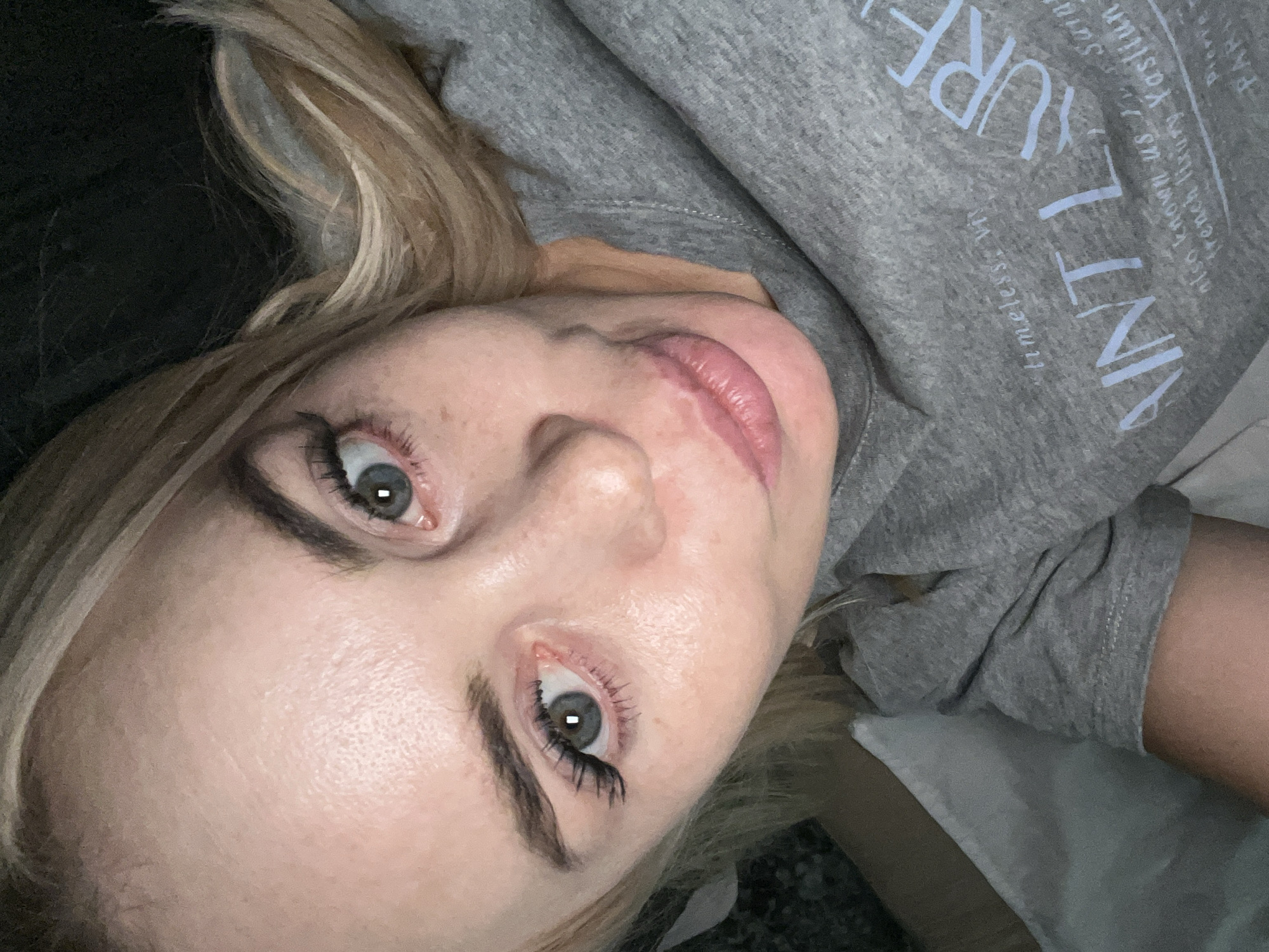 A close-up, high-angle, upside-down shot shows a person lying down, with their head tilted. They have fair skin, blonde hair with highlights, and blue eyes. The person is wearing a grey sweatshirt with light blue text on it. The text is partially visible and appears to read "AINT LAURI" and other words that are difficult to decipher. The person's makeup includes mascara and what appears to be lip gloss. The background is dark and out of focus, suggesting the setting might be indoors. The lighting is soft, and the overall mood is casual and relaxed.