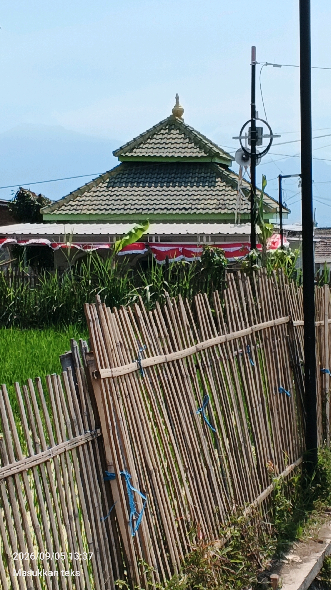 The image shows a mosque with a green roof and a tiered design, featuring a golden finial at its peak. The mosque is surrounded by tall green grass and what appears to be a rice paddy field in the foreground. A fence made of bamboo poles runs diagonally across the lower portion of the image. Red and white banners are draped along the front of the mosque building, suggesting a celebratory occasion. A black pole with what looks like a speaker or antenna is visible to the right of the mosque. The sky is a clear, pale blue, with a hint of distant mountains. The date stamp "2026/09/05 13:37" is visible in the bottom left corner. The scene is located in Jeru, Indonesia.