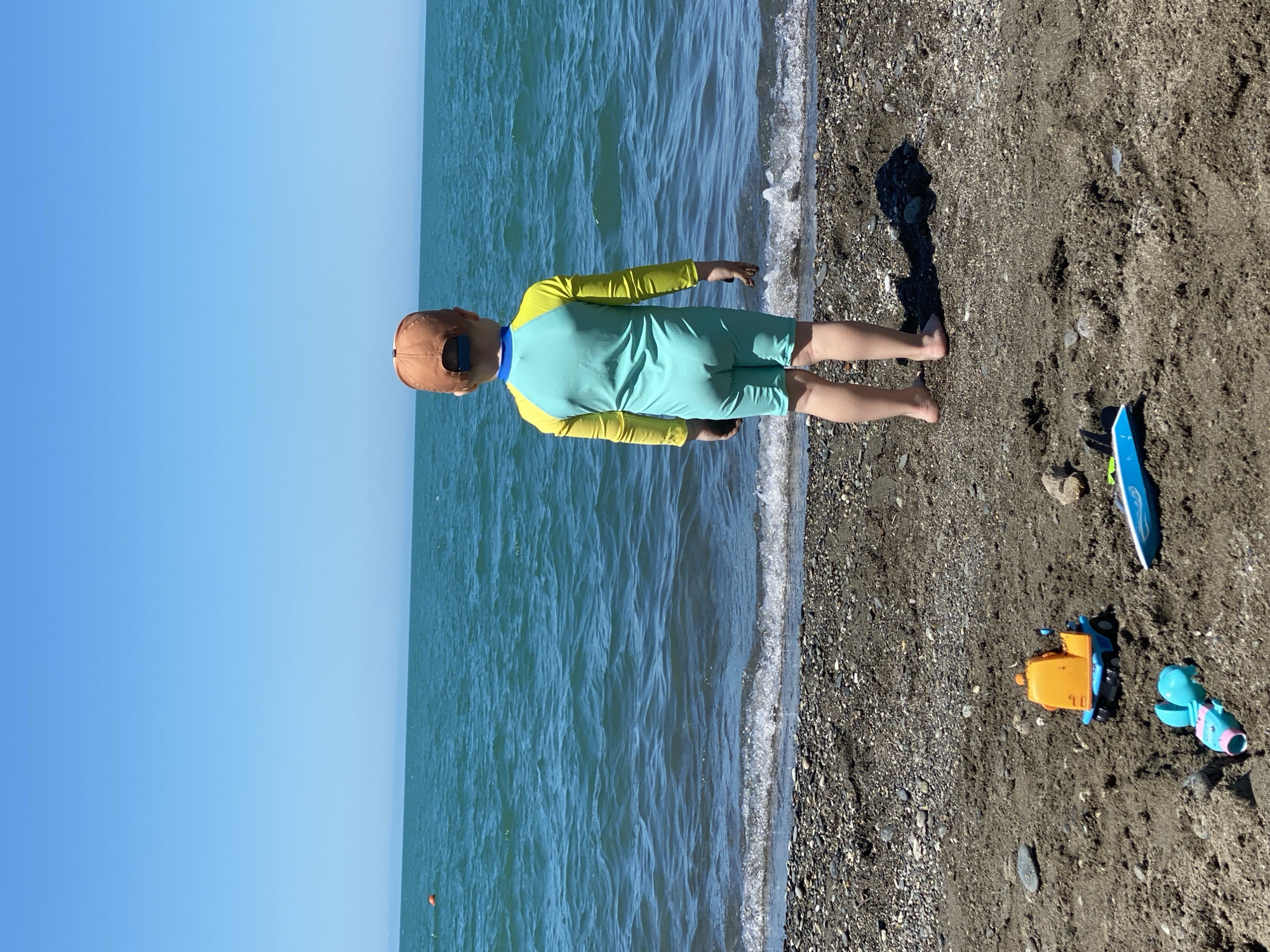 This image captures a young child standing on a pebble and sand beach in Sochi, Russia, facing the Black Sea. The child, viewed from behind, is wearing a light teal full-body swimsuit with bright yellow long sleeves and a brown baseball cap. Their bare feet are positioned at the edge of the water where small, foamy waves are breaking. Dark sand smudges are visible on their hands. The beach consists of dark grey and brown sand intermixed with numerous small pebbles. In the foreground, several beach toys are partially buried in the sand: an orange and blue toy dump truck, a light blue toy boat, and a teal and pink plastic toy. The sea beyond is a calm expanse of turquoise-green water with subtle ripples, and a single orange buoy is visible floating in the upper left section of the water. The horizon line is clear and level, separating the sea from a uniformly bright, cloudless blue sky. The child casts a distinct shadow to their right, indicating a bright sunny day.