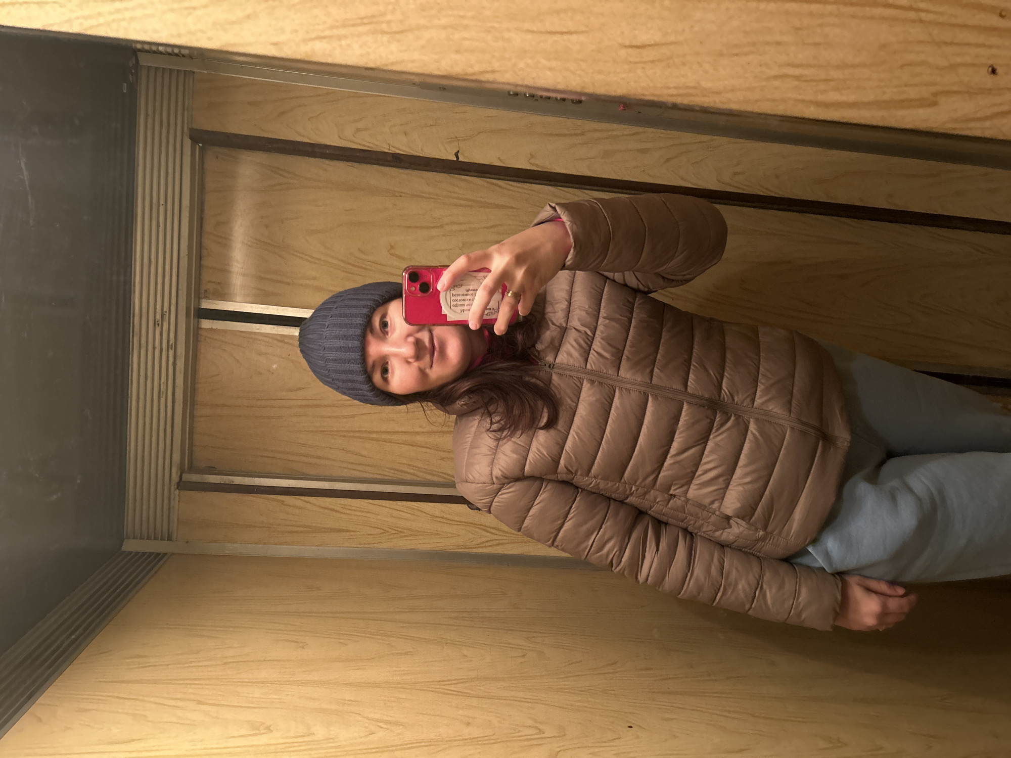A woman takes a selfie in an elevator. She is wearing a blue beanie, a brown puffer jacket, and light blue sweatpants. She holds a red phone in her hand. The elevator has wood-paneled walls and a dark grey door. The location is unknown.