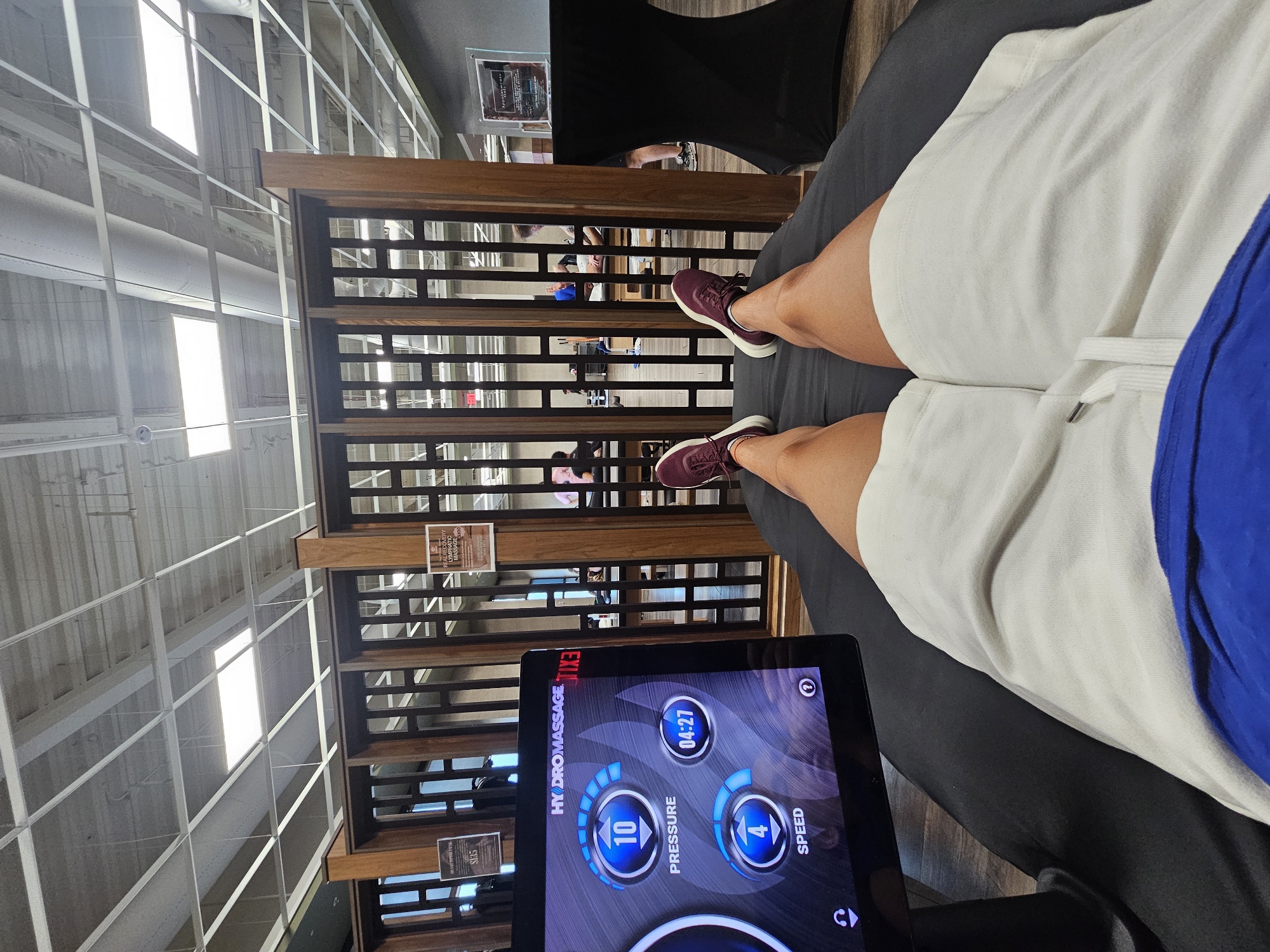 A first-person point-of-view photograph shows a person lying on a black massage bed, wearing white shorts and maroon sneakers, while operating a massage machine. The machine’s tablet screen displays the brand “HYDRO MASSAGE” and the massage settings, indicating a pressure level of 10 and a speed of 4. The background reveals a wooden partition with horizontal slats, and through these slats, other people can be seen in the background. The environment appears to be indoors. Based on the visible signage “REAL RECOVERY LYMPHATIC MASSAGE, MD,” it is likely that the location is in Miami, United States of America.