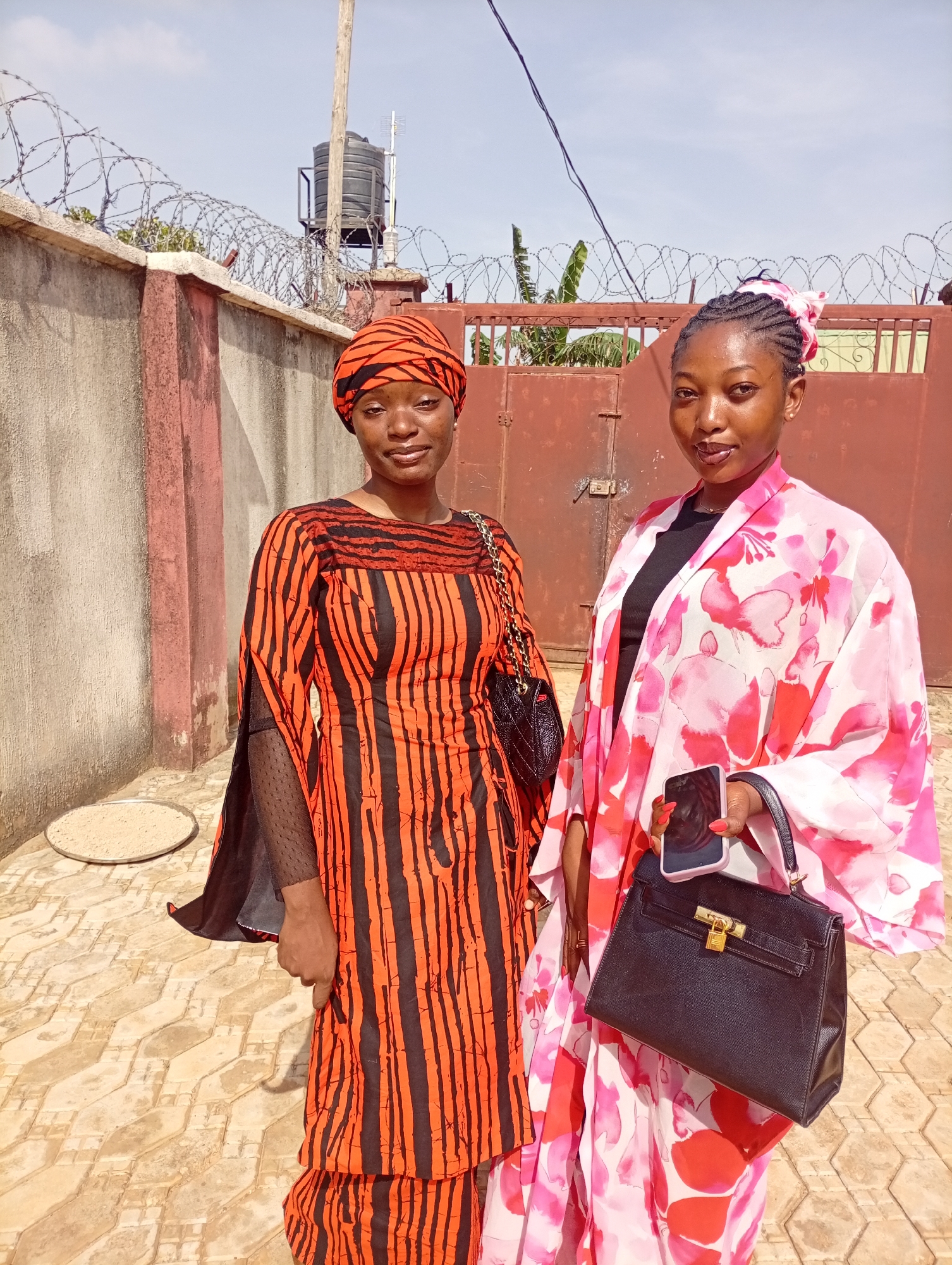 Two women stand side by side in an outdoor setting. The woman on the left wears an orange and black striped dress with a matching head covering. She has a black purse hanging from her shoulder. The woman on the right is dressed in a pink floral kimono-style garment over a black top. She is holding a black handbag in her left hand and a mobile phone in her right hand. The background features a concrete wall topped with barbed wire and a tall metal gate. There is a water tank visible above the wall. The ground is paved with patterned tiles. The location is in the city of Unknown, Unknown.