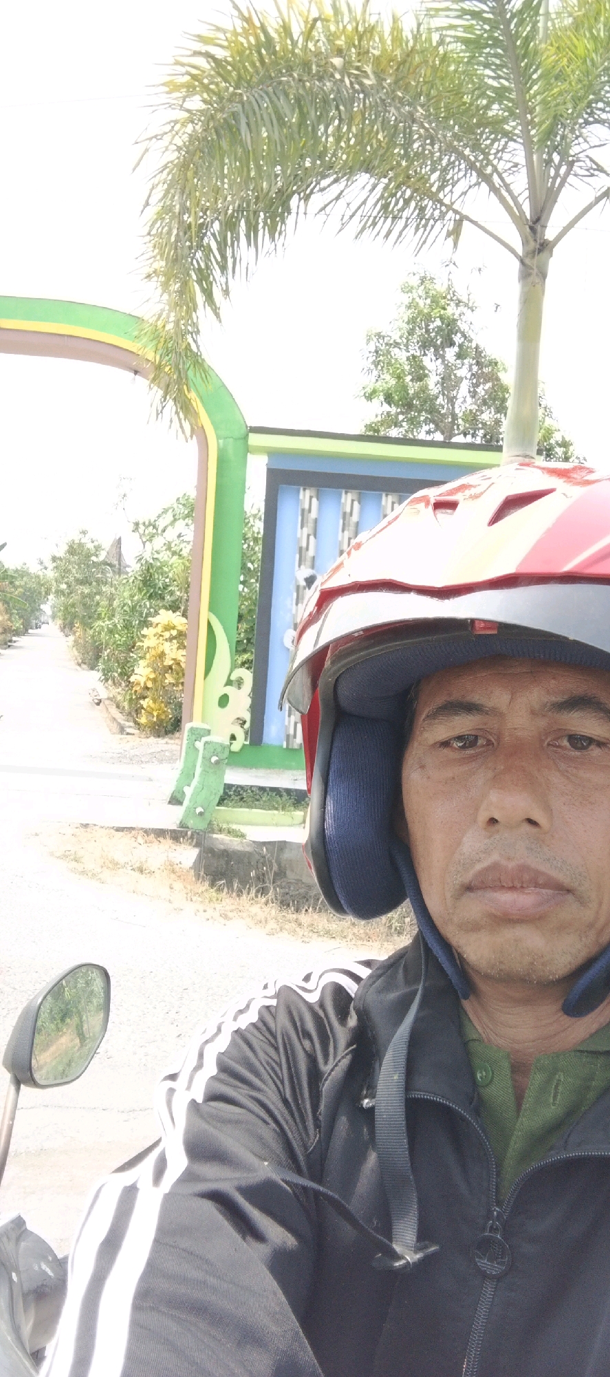 In Guci, Indonesia, a man wearing a red helmet and a black jacket with white stripes is taking a selfie while riding a motorcycle. He is looking at the camera with a neutral expression. The background shows a street lined with trees and a colorful archway. The image is taken from a low angle, looking up at the man.