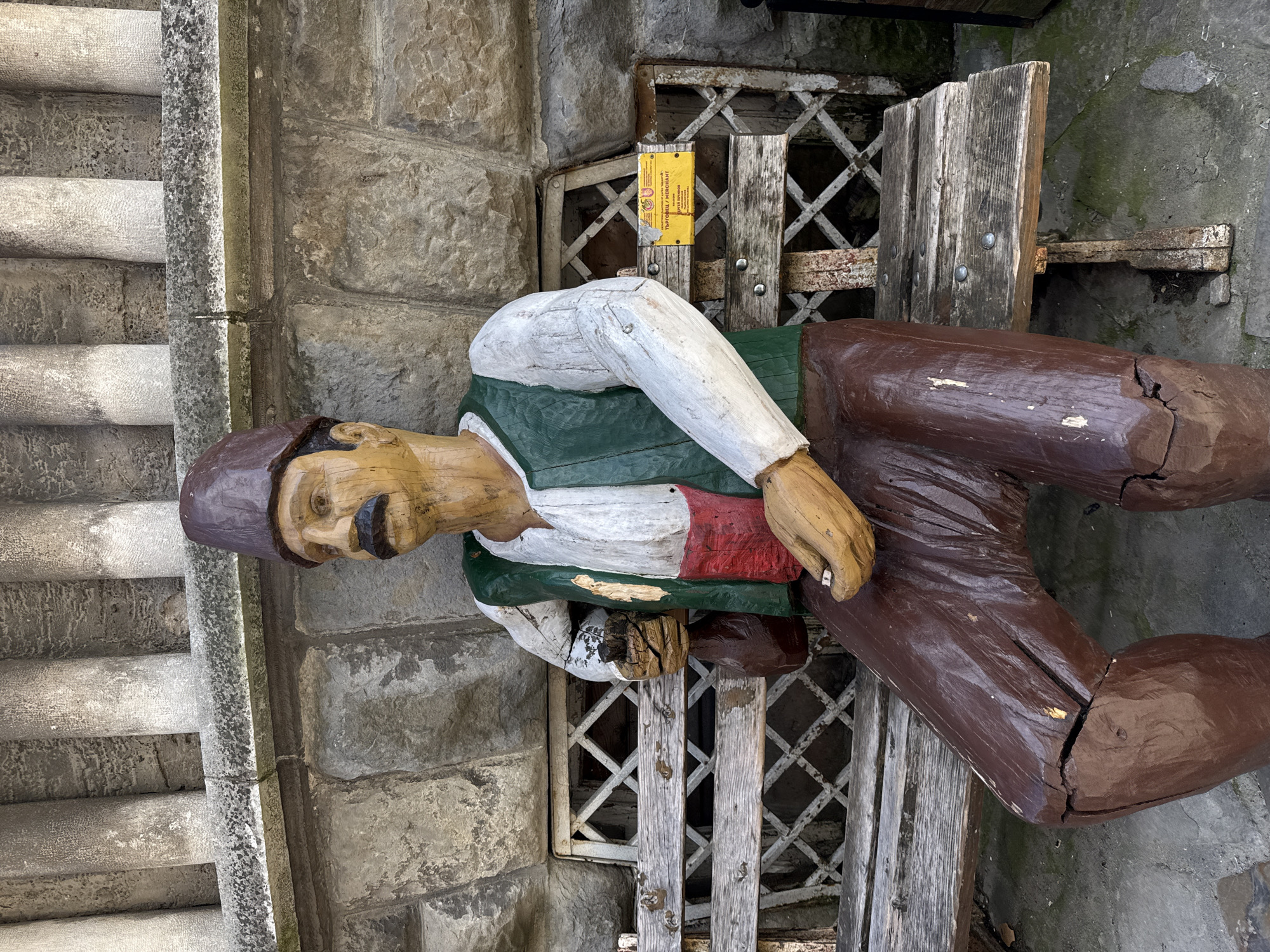 A carved wooden statue of a seated man rests against a stone wall in Veliko Tarnovo, Bulgaria. The statue is painted with a brown cap, a yellow face with a black mustache, a white shirt with a green vest and red sash, and brown pants. The statue is sitting on a wooden bench in front of a metal grill. To the left of the statue are stone stairs. A yellow sign is visible behind the statue. The statue appears to be weathered and cracked. The scene is in daylight.