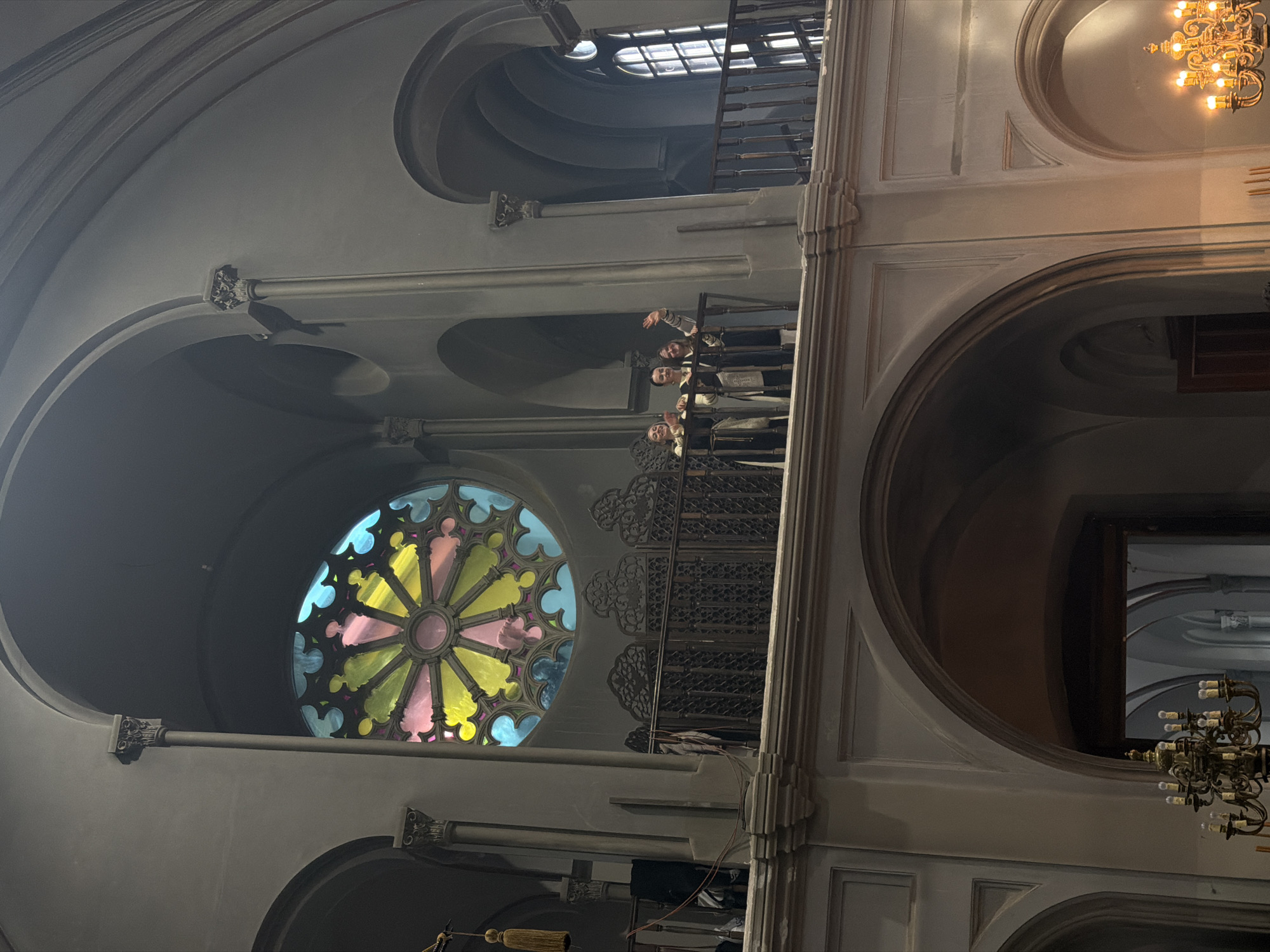 The image captures the grand interior of a building in Saint Petersburg, Russia, likely a church or concert hall, featuring ornate architecture and multiple levels. The perspective is looking upwards towards a high balcony and ceiling.
The main subjects are several people standing on a metallic-railed balcony or choir loft on the left side of the frame. They appear to be looking down towards the viewer or the main floor. One person in the foreground of the group is waving, while others are also observing. They are dressed in formal attire, possibly choir robes, suggesting they might be performers or participants in a service or event.
Prominently featured is a large, circular stained-glass rose window located below the balcony. It displays vibrant colors, including yellow, pink, blue, and green, with light filtering through. Above this, and higher up in the building, are arched ceilings and an upper window with a grid pattern.
Architectural details abound, including grand arches, fluted columns with ornate capitals, and decorative mouldings on the walls and ceilings. To the right, an elaborate golden chandelier with multiple bulbs illuminates a section of the wall. Another decorative ceiling element with a circular motif is also visible on the right.
Notable details include a small, dark bird perched on a horizontal rod or decorative element within an arch on the upper left, an unexpected presence in the grand interior. The lighting is dim overall, with concentrated light from the stained-glass window and chandeliers, indicating either an interior shielded from direct sunlight or a low-light setting. Cables or wires are visible hanging near the balcony, possibly for lighting or audio equipment. The building exhibits a sense of history and grandeur. No visible text is present in the image.