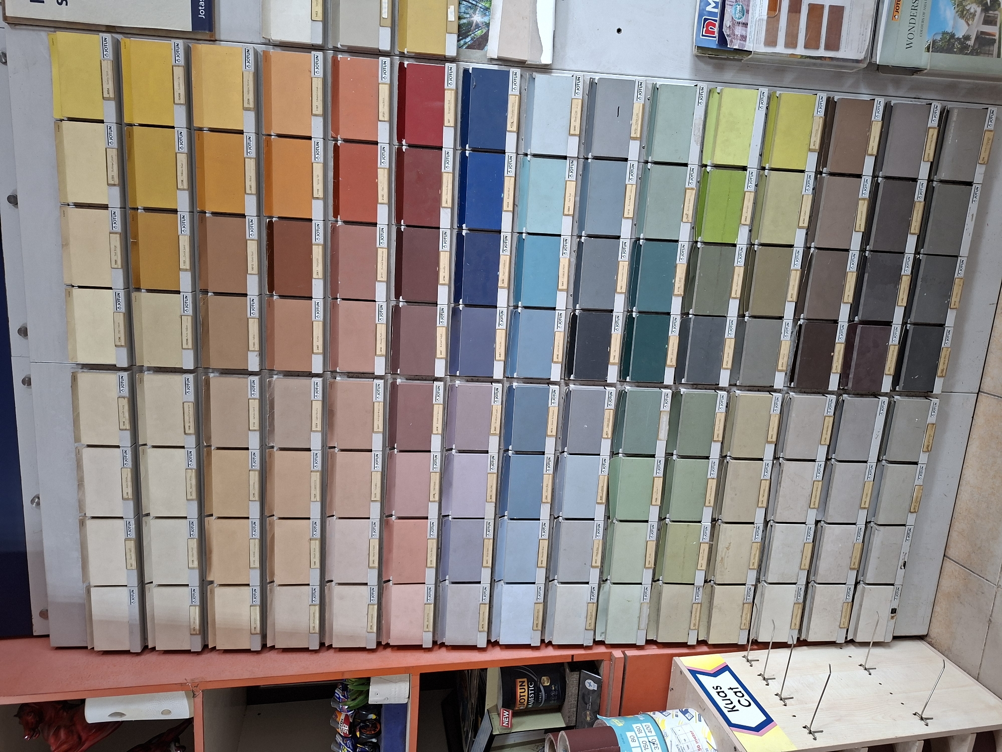 The image shows a wall-mounted display of paint color swatches, likely in a hardware or paint store in Lhokseumawe, Indonesia. The display is organized with horizontal rows and vertical columns of rectangular color samples. Each sample has a label, the brand name "Jotun" is visible on many. The color palette spans a wide range, including yellows, oranges, reds, blues, greens, grays, and browns.
Below the paint swatches is an orange shelf. On the shelf, there are various items, including cans and a box with the words "Kuas Cat" (Paint Brush). Other objects on the shelf suggest this is a retail environment.