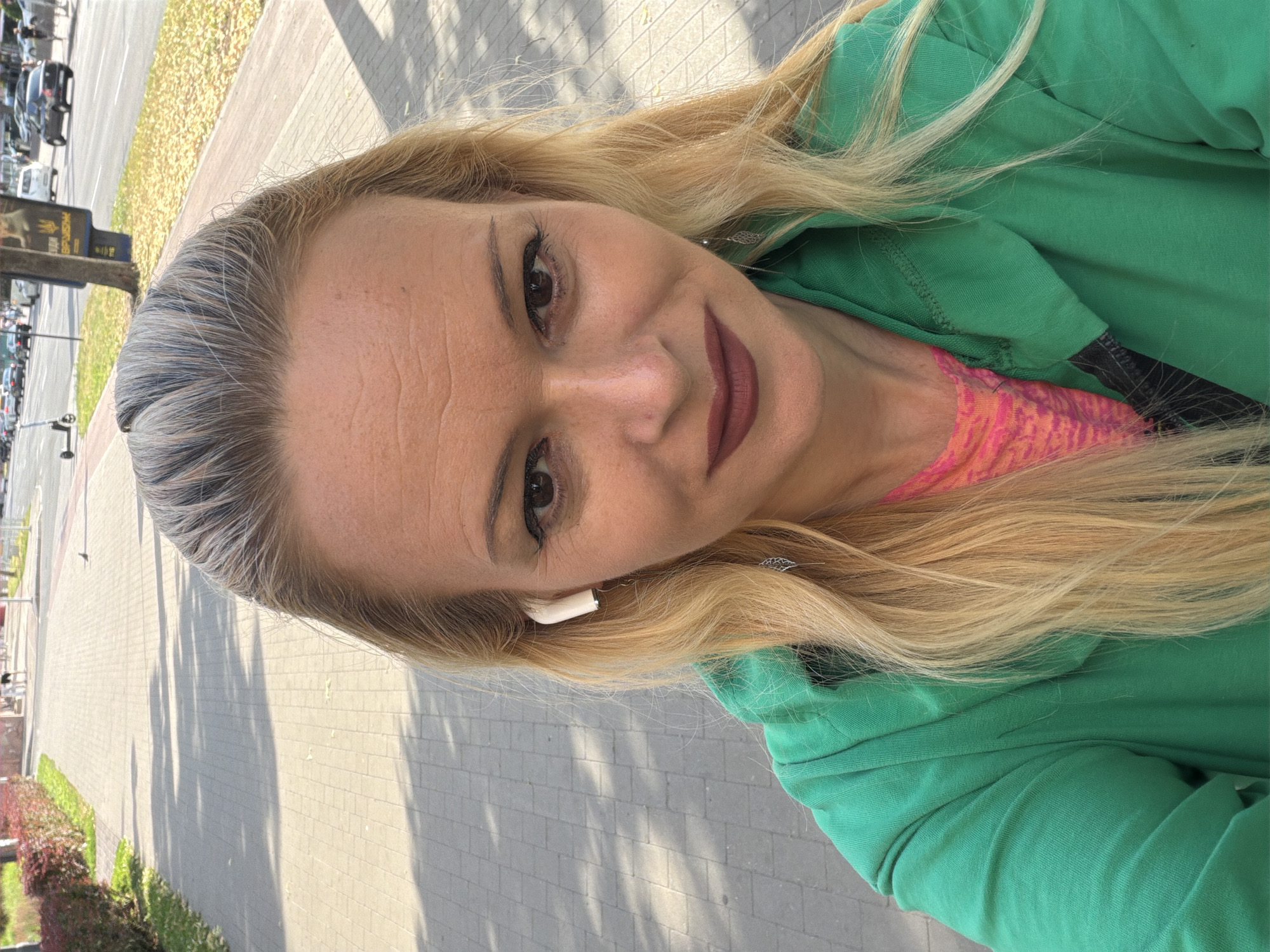 A woman with blonde hair and fair skin is taking a selfie on a paved sidewalk in Chernihiv, Ukraine. She is wearing a bright green hooded jacket and a pink shirt. An earbud is visible in her right ear. The background shows a street with cars, a sidewalk, a grassy area with bushes, and a tree with a blue sign. The sun is casting shadows on the sidewalk.