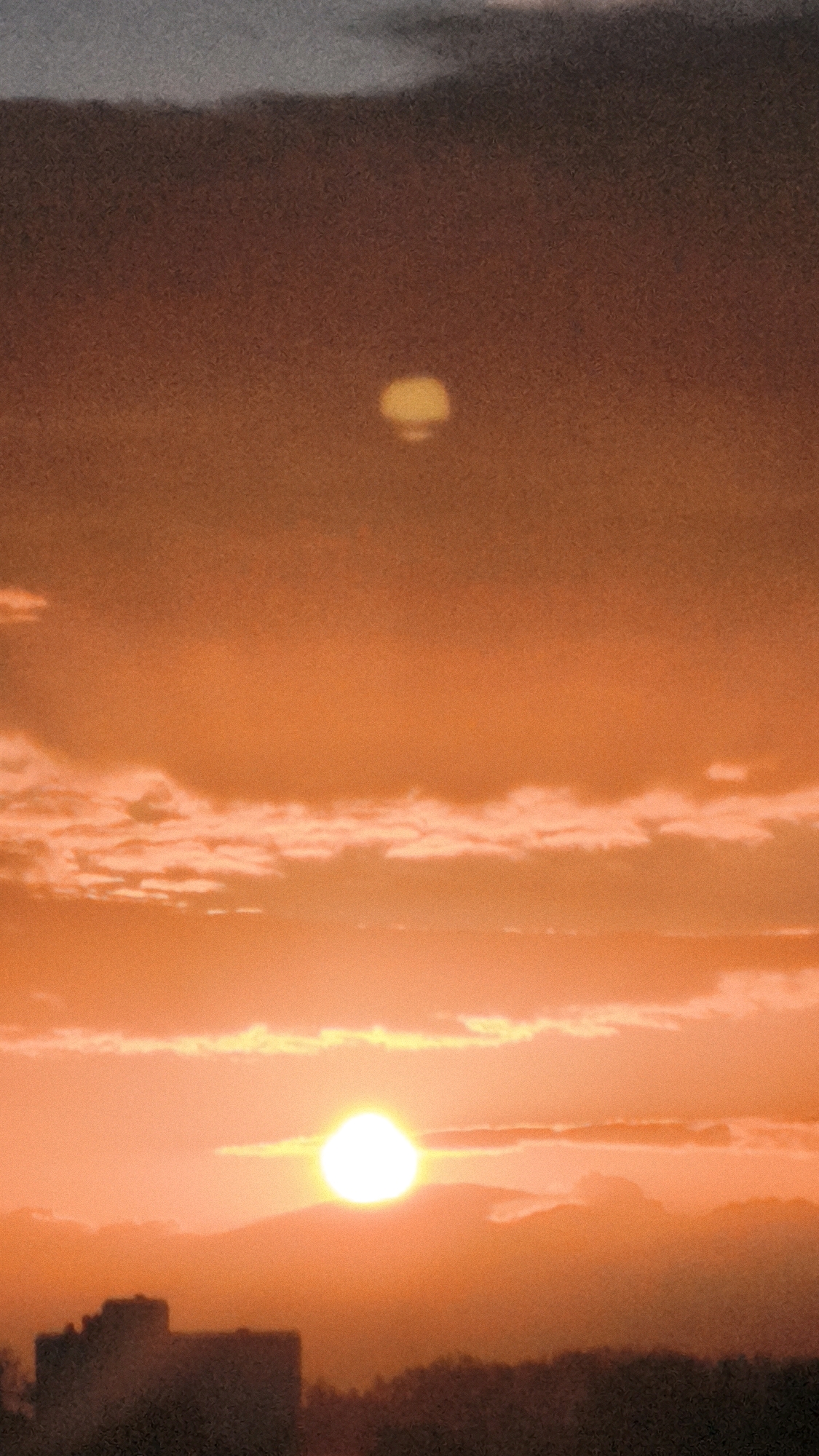 The image depicts a sunset over the city of Worms, Germany. The sky is filled with shades of orange and yellow, with some scattered clouds. In the foreground, silhouetted buildings and trees are visible against the vibrant sky. The sun, a bright orb, is setting near the horizon, casting a warm glow over the scene. The overall impression is one of a peaceful and beautiful end to the day.