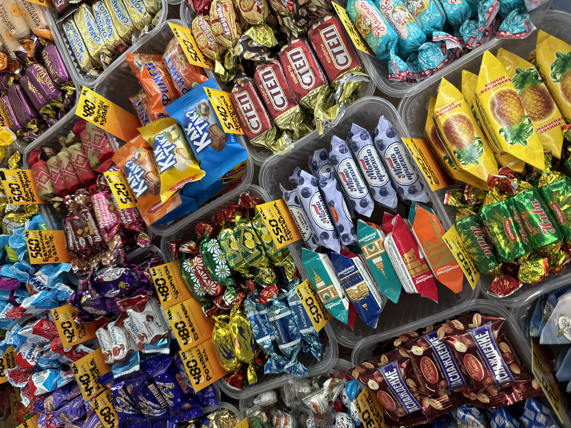 The image shows a diverse selection of unwrapped candies and chocolates displayed in clear plastic containers, likely in a retail setting such as a market or confectionery shop in Rusanovo, Russia. Each container holds a different type of confection, with various brands and flavors represented. Small yellow price tags are affixed to many of the candy varieties, indicating the price per unit weight. The candies range from chocolate bars and caramel candies to fruit-flavored sweets and wafer-based confections. There are no people visible in the shot. The overall impression is of a colorful and abundant assortment of sweets available for purchase.