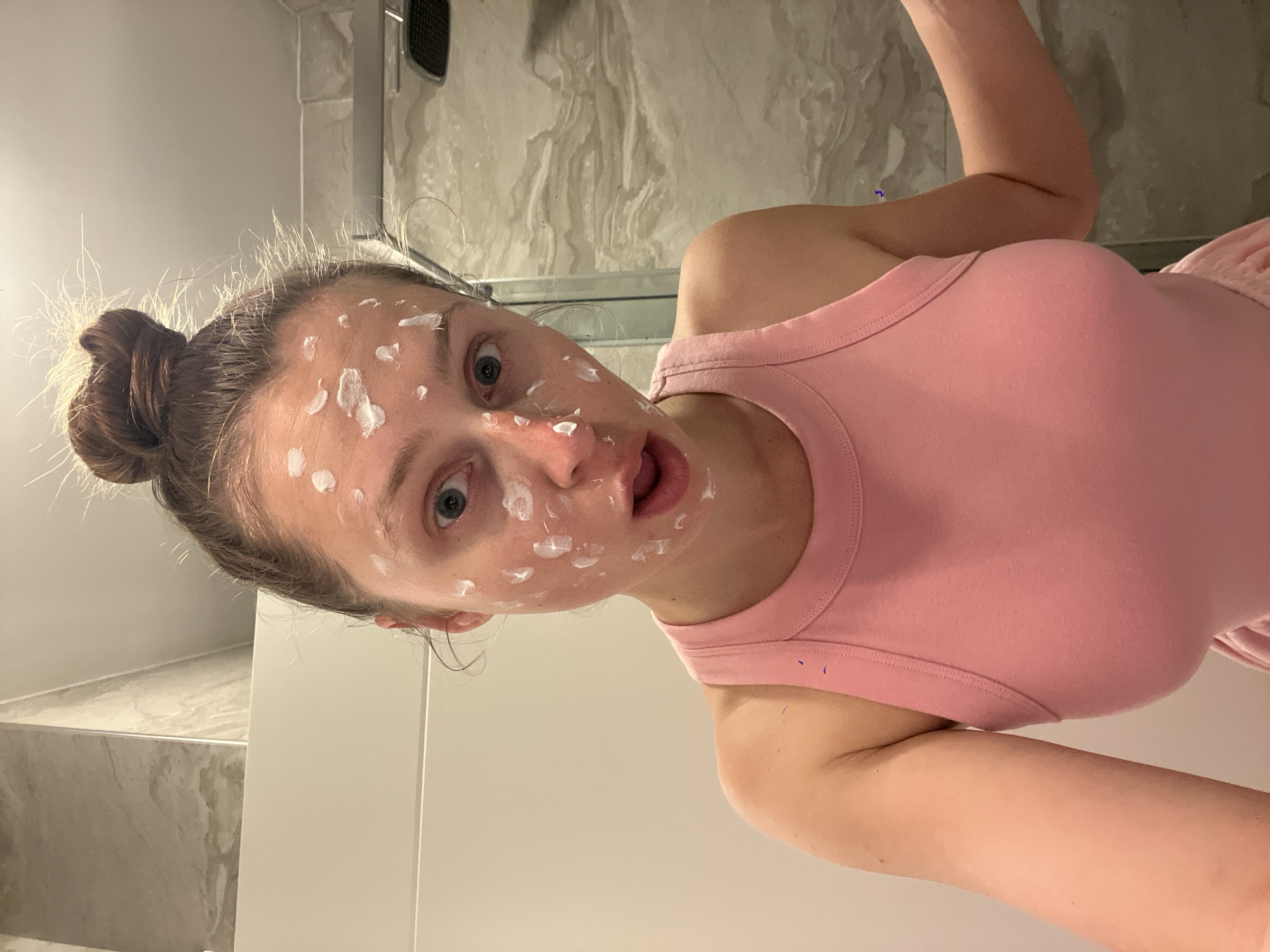 A woman with her hair in a bun is posing in what appears to be a bathroom. She is wearing a pink tank top and has white spots on her face, possibly from a cosmetic treatment. The background shows a marble-patterned wall, a shower fixture, and a white cabinet. There is no indication this image was taken in Rafailovići, Montenegro.