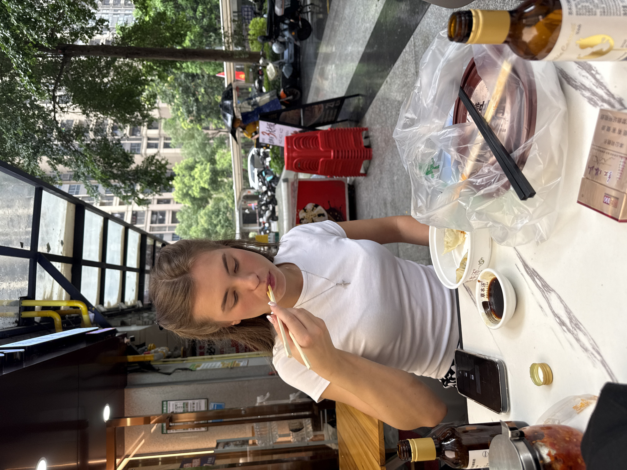 This media file is a photograph taken outdoors in Haizhu District, China, likely during the day given the natural light. The scene appears to be a street-side dining area with a woman seated at a table.
The primary subject is a young woman with blonde hair, wearing a white t-shirt and a silver necklace. She is actively eating, holding chopsticks to her mouth. Her eyes are closed, suggesting she might be savoring the food or simply in the moment.
The table is set for a meal. Visible objects include a white bowl with what appears to be dumplings or similar food items, a small dish of dark dipping sauce, a mobile phone, a plastic bag containing more food items and chopsticks, and a brown glass bottle with a yellow label, possibly containing a beverage. There is also a gold-colored ring on the table. Further back, there are stacks of red plastic chairs, more tables, and umbrellas, hinting at a casual outdoor eatery.
In the background, the scene transitions to a street with various vehicles, including motorcycles, and buildings. Trees with lush green foliage are also present, indicating a possibly warm day. The overall atmosphere seems relaxed and everyday.
Visible text in the image includes "草本汤" (herbal soup) and "红塔山" (Hongtashan, a brand of cigarettes), along with a health warning about smoking. The Chinese characters suggest the location is likely in China.