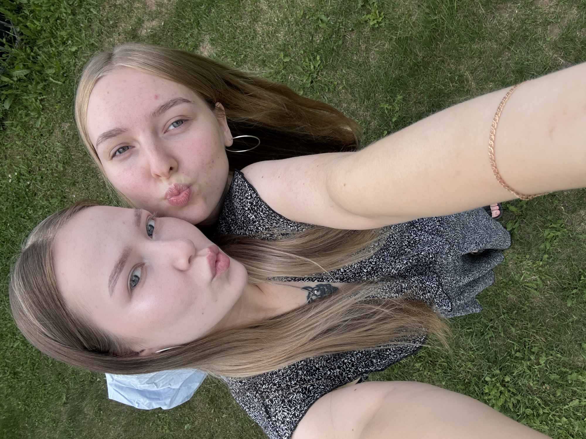 Two young women are taking a selfie outdoors on a grassy area, with lush green grass filling the background. The woman on the left has long, straight, light brown hair and is wearing a black and white patterned dress and large hoop earrings. She has a tattoo on her chest. The woman on the right, with blonde hair, is leaning in and puckering her lips towards the camera. She is also wearing hoop earrings. The lighting suggests it is daytime. The location is identified as Zhigalki, Russia.