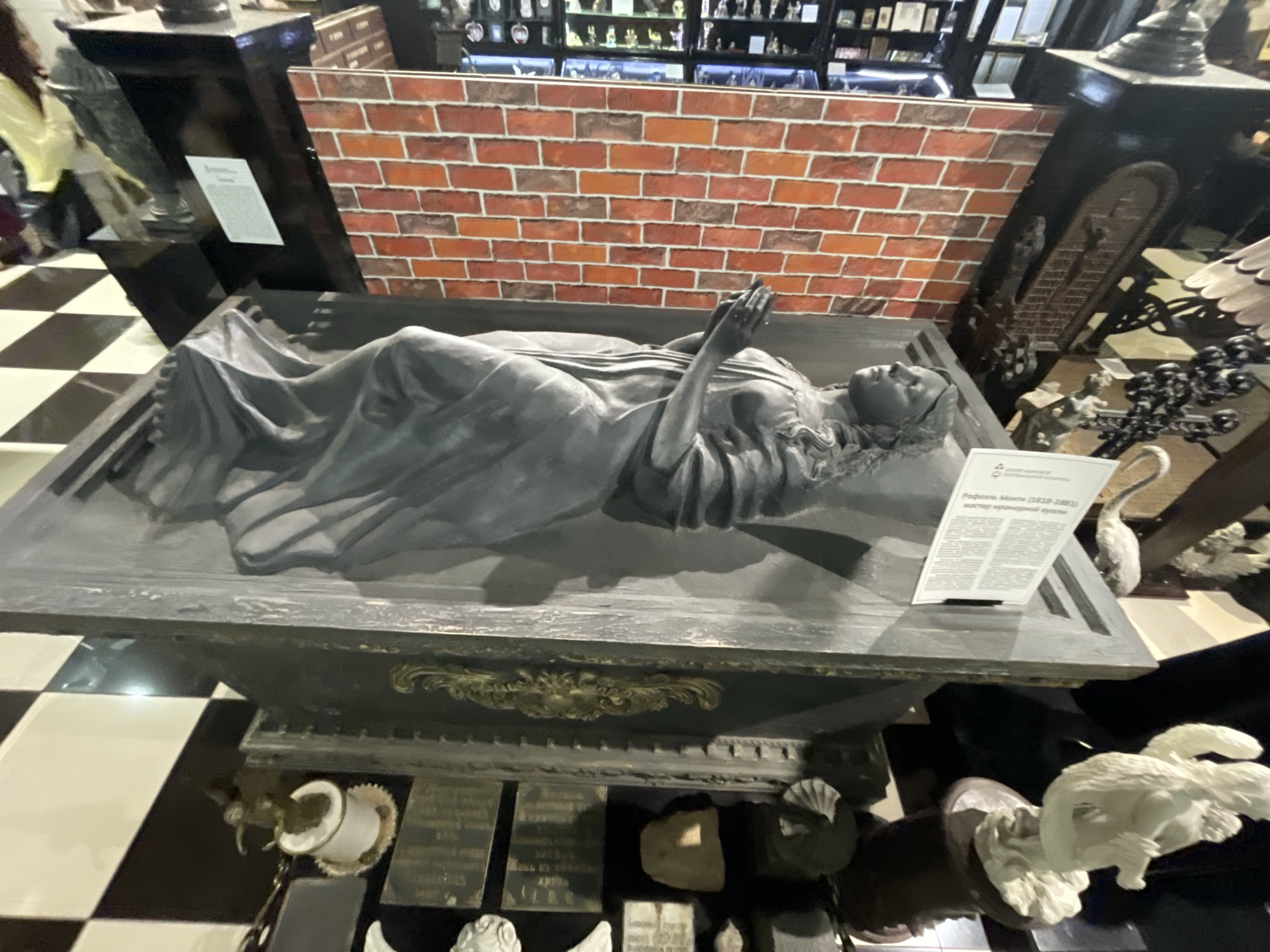 The image depicts a museum exhibit featuring a sculpture of a reclining figure, possibly a representation of death or a saint, lying in a sarcophagus-like structure. The sculpture is draped in flowing fabric, and the figure's hands are clasped together in prayer. Behind the sculpture is a brick wall backdrop. The scene is set in what appears to be a dimly lit museum with a checkered floor. Several other exhibits and decorative elements are visible in the background and foreground, including small figurines and ornamental pieces. A plaque with text is placed next to the sculpture, providing information about the artwork and artist, identified as "Rafal Monti (1818-1882), master of monumental sculpture." The text on the plaque is in Russian. There are no people clearly visible interacting with the exhibit. The image is a wide shot, capturing the entire display. The overall atmosphere is somber and artistic, typical of a museum setting. The location is confirmed as the Museum of History and Local Lore in Voskhod, Russia, based on the information on the plaque.