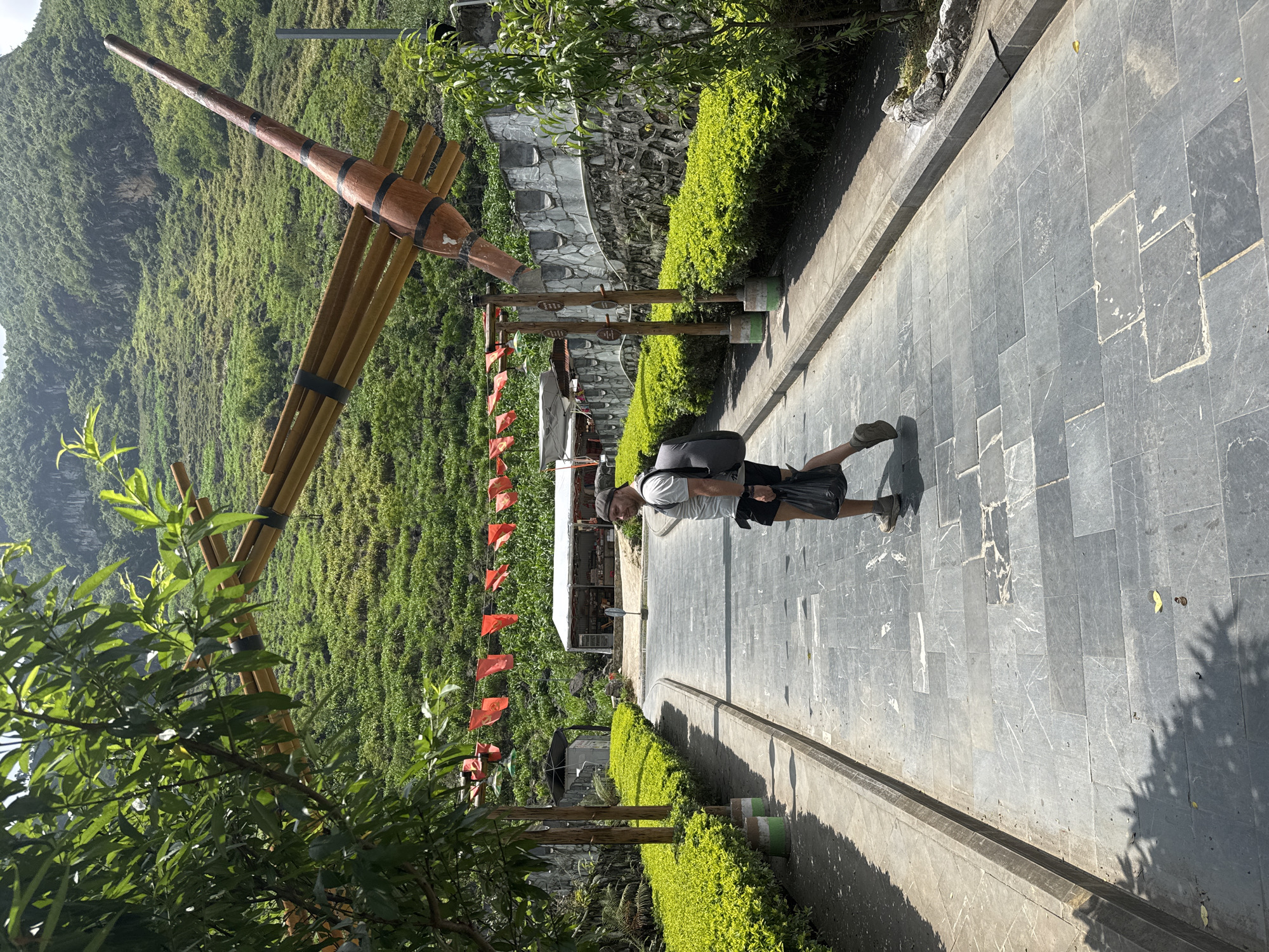 A man stands on a gray stone pathway in Mèo Vạc Commune, Vietnam. The path is bordered by manicured hedges and leads towards a small structure with a white roof. In the background, a large, decorative bamboo structure resembling a musical instrument stands against a lush green hillside. Red flags hang from the structure's support beams. The man is wearing a white shirt, dark shorts, a hat, and a backpack. He appears to be walking along the path. The environment is bright and sunny.