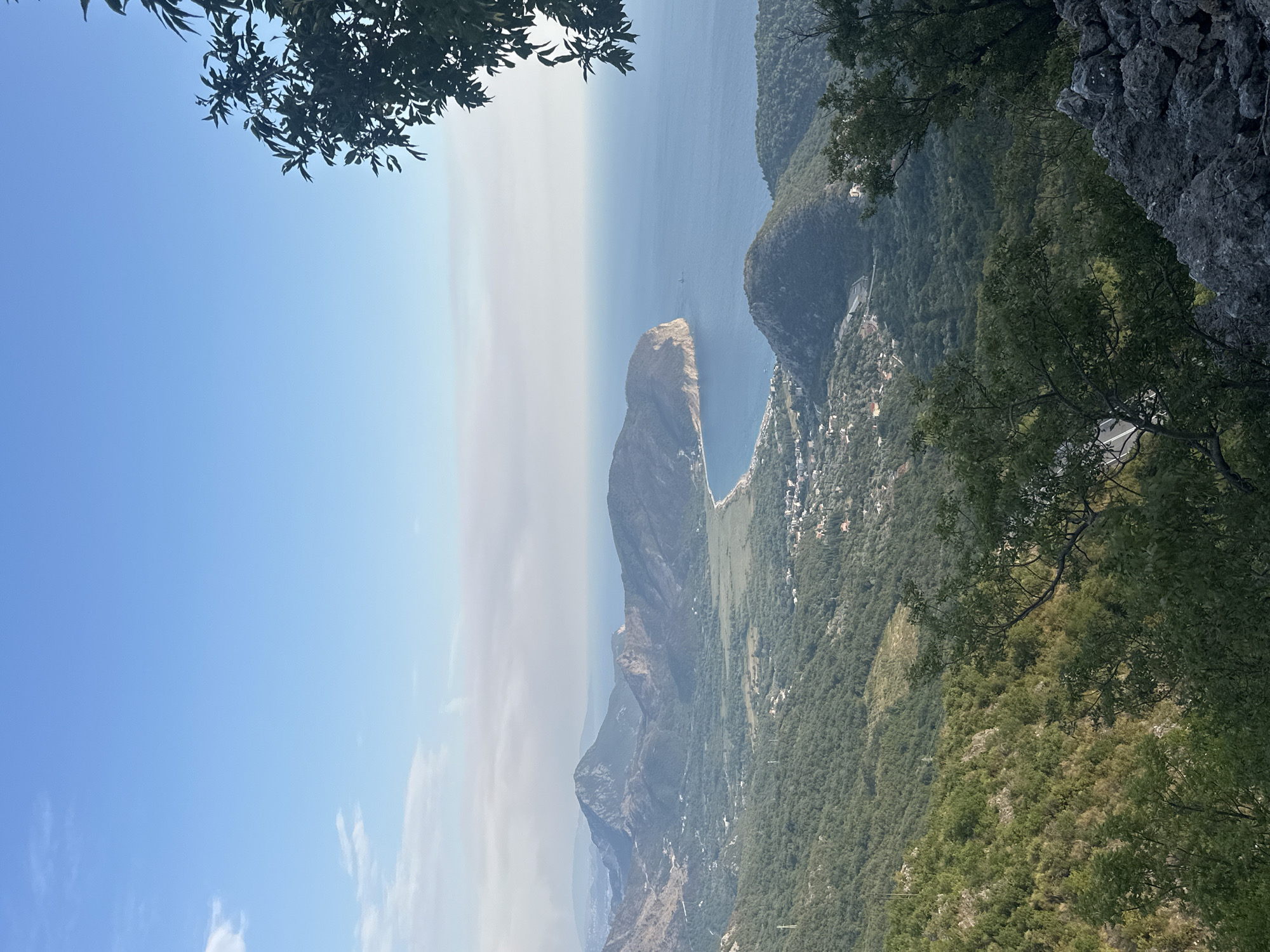 This image, captured from a high vantage point in Novoselje, Montenegro, offers a panoramic view of a coastal landscape. In the foreground, rugged grey rocks and dark green foliage of trees frame the scene. Beyond this, a verdant valley slopes down towards the sea, dotted with scattered houses and buildings that comprise the settlement of Novoselje. The valley opens up to a crescent-shaped bay with a sandy beach, leading to the deep blue Adriatic Sea. In the distance, a series of majestic, undulating mountains rise, their peaks softened by a hazy atmosphere. The sky is a clear, pale blue with a few wispy clouds. A lone sailboat is visible on the water in the mid-ground. The overall impression is one of serene natural beauty and a peaceful, coastal settlement.