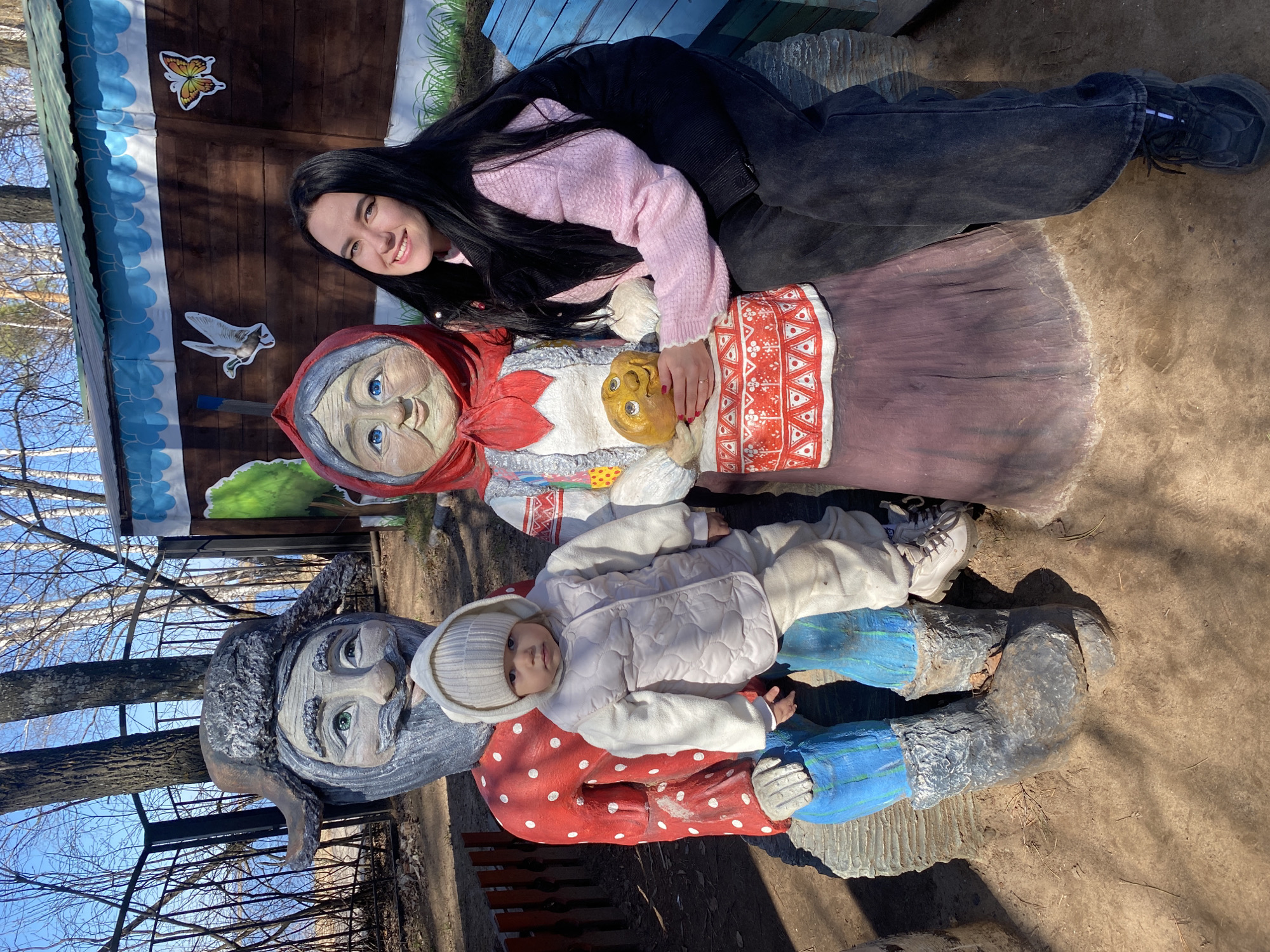 A woman with long black hair is lying on a painted statue of an old woman. A child in a white jacket and hat lies on top of a statue of a man with a beard. The location is the City of Chaikovsky, Russia. A wooden building with painted images is visible in the background. Trees are visible in the distance. The ground is dirt and grass.
