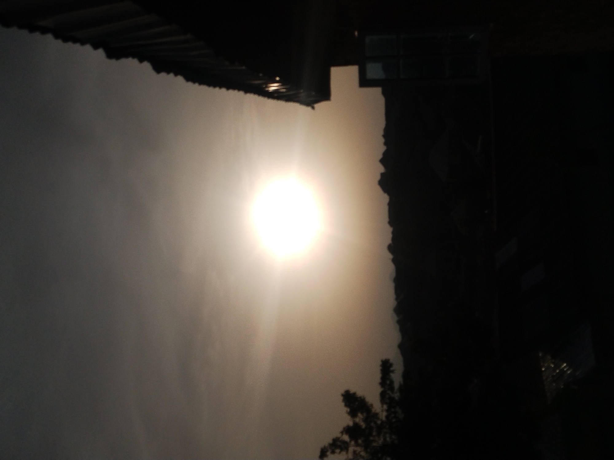 The image depicts a landscape under a bright sun. The foreground shows a sky obscured by a bright light source. In the upper portion of the image, the dark silhouette of a rooftop structure is visible. The lower right corner contains a dark area with a tree. Given the presence of rooftop structures and trees, the image may depict an urban scene. It is not possible to confirm the location or subjects in the image. There are no identifiable people, actions, or interactions.